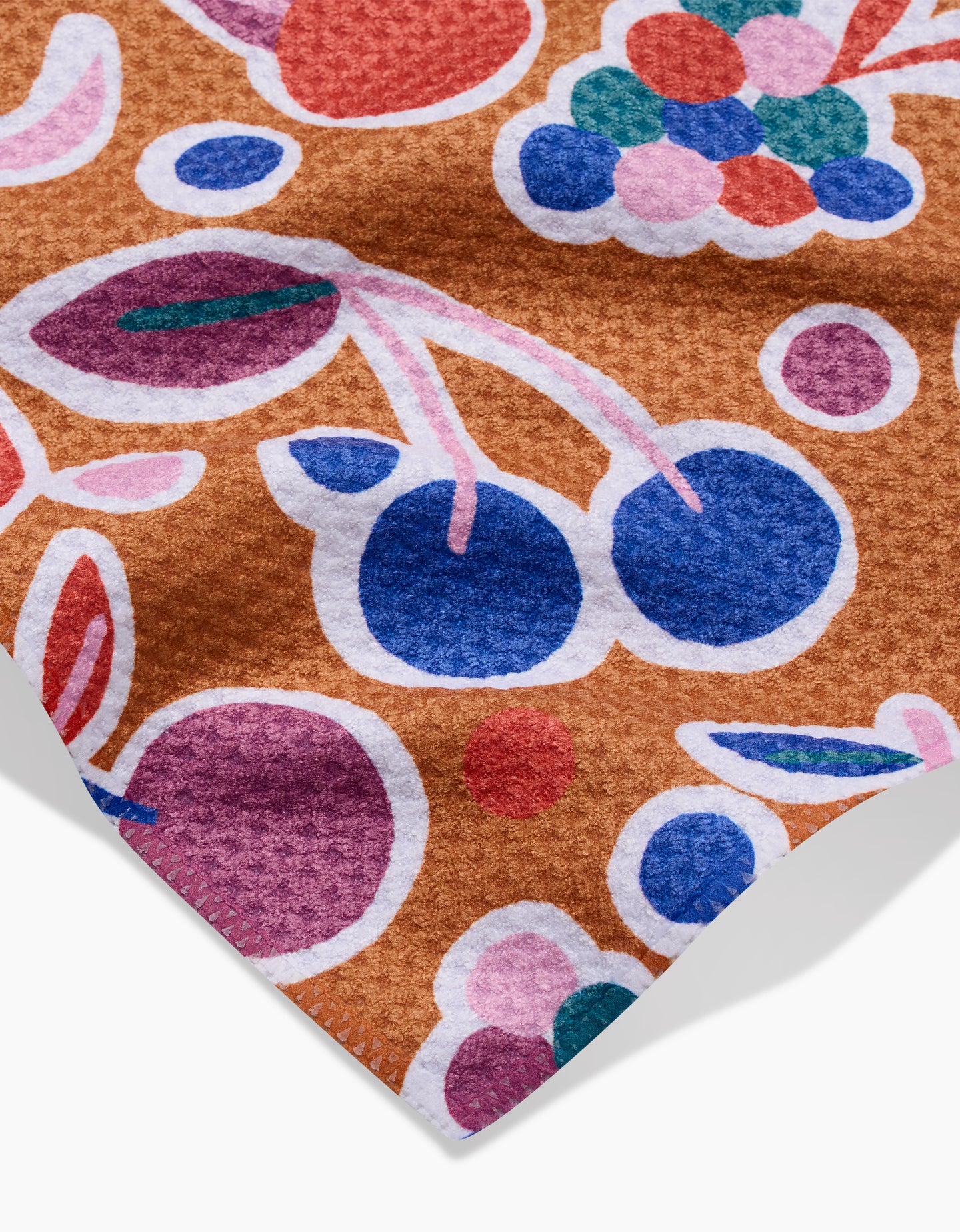 A close-up of the Claire Iglesias Fruits Party Bar Towel shows a colorful, abstract fruit pattern with blue, pink, and purple shapes on a textured brown Geoweave fabric.