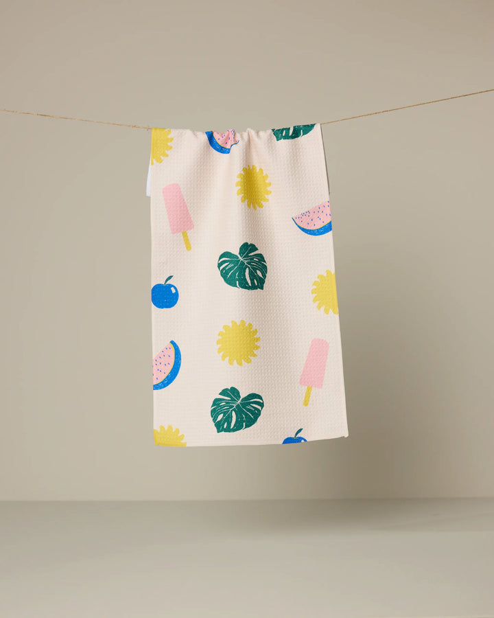 The Geometry Fruity Pop kitchen towel, featuring a vibrant print of watermelon slices, ice cream popsicles, flowers, cherries, and monstera leaves on beige fabric, hangs on a clothesline against a plain backdrop.