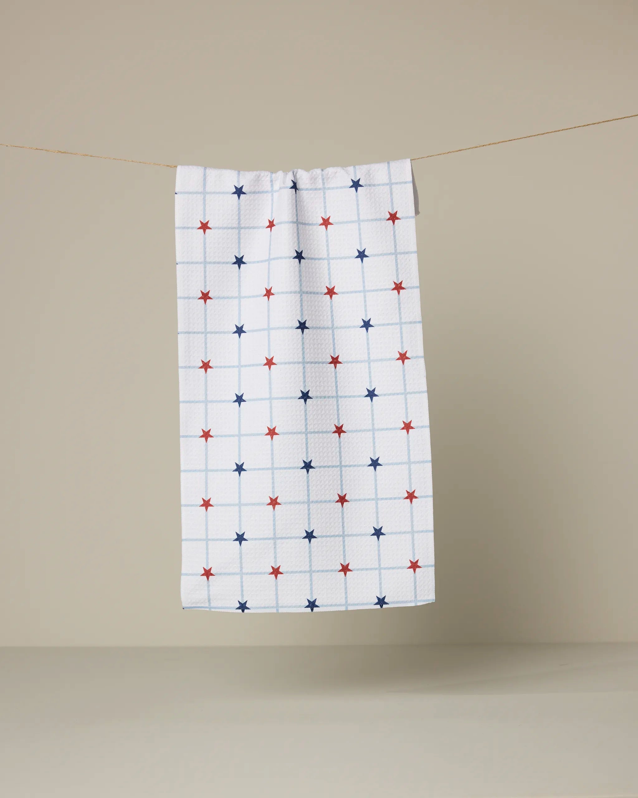 The Garden Stars bar towel by Geometry, featuring a blue grid and red and blue stars on white Geoweave fabric, hangs on a clothesline against a plain beige background.