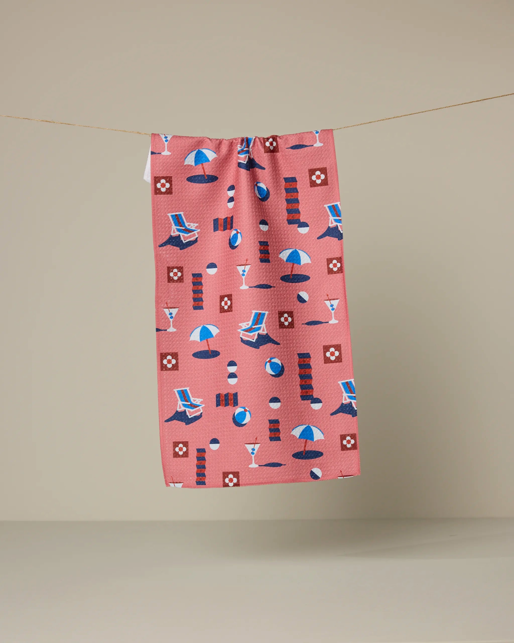 The Geometry Geo Beach Day towel, in pink and ultra-absorbent, hangs on a beige background, showcasing playful beach-themed prints like umbrellas, lounge chairs, beach balls, drinks, lifebuoys, and striped towels.