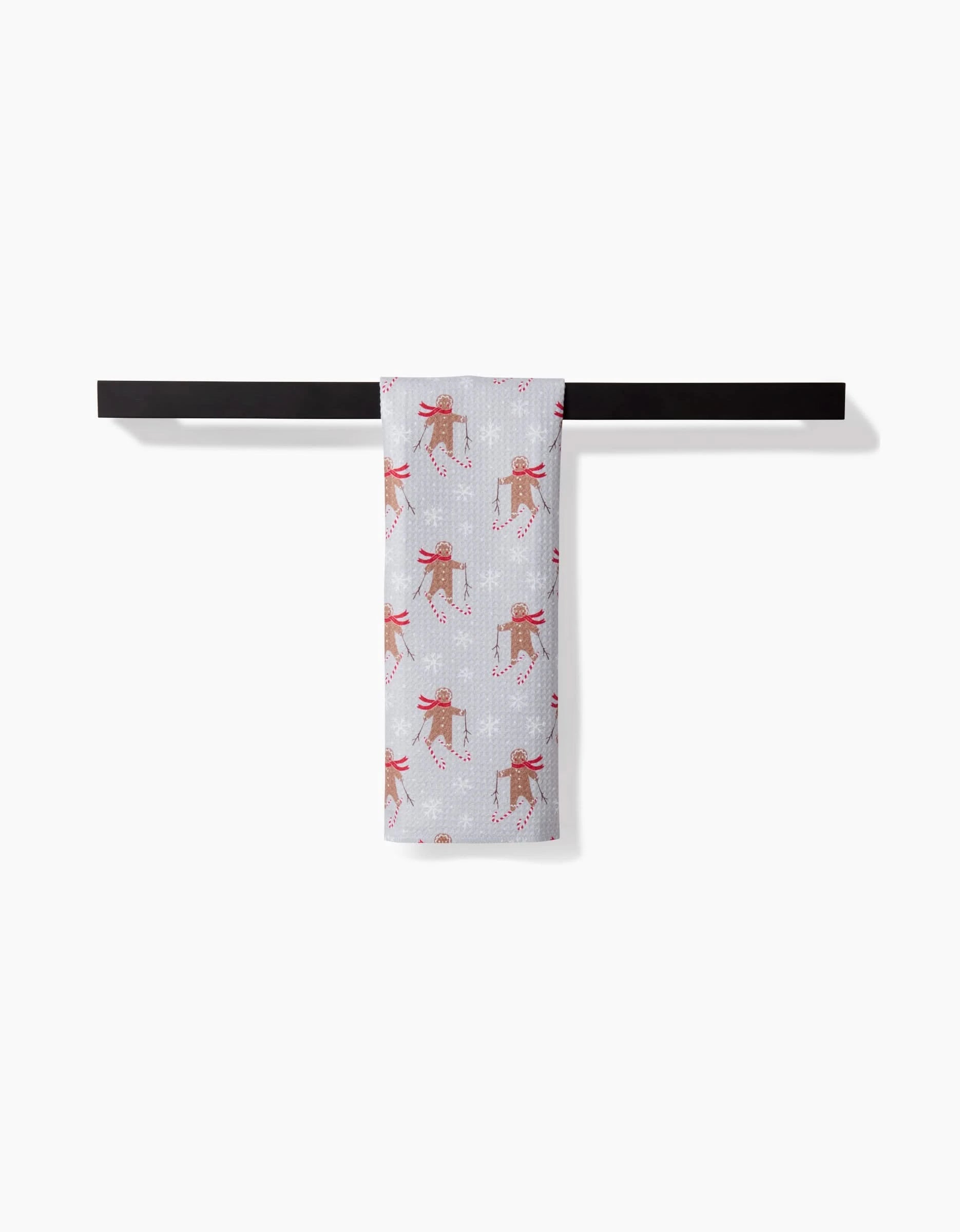 A Gingerbread Skiers kitchen towel by Roxy Marj featuring a festive design hangs on a black horizontal bar against a plain white background.