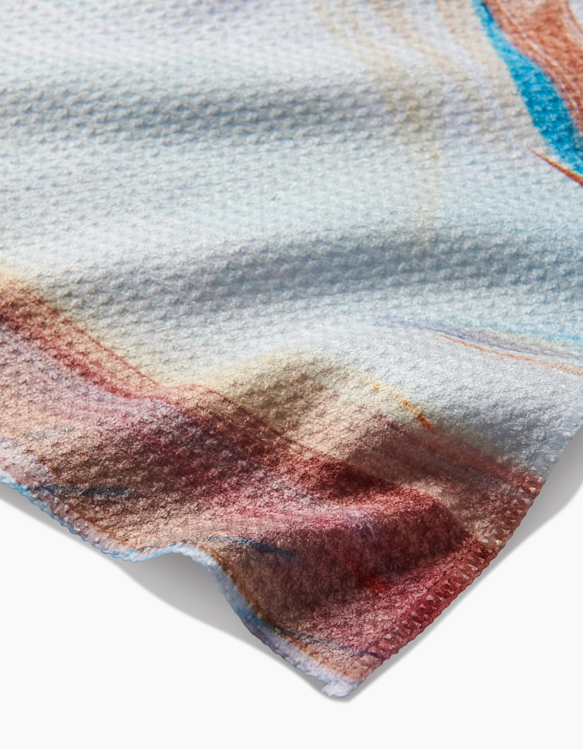 A close-up of the Kelly Peters Golden Hour in the Rockies Bar Towel shows its textured Geoweave fabric with abstract blue, beige, and reddish-brown patterns. The towels edge casts a soft shadow on a white background.