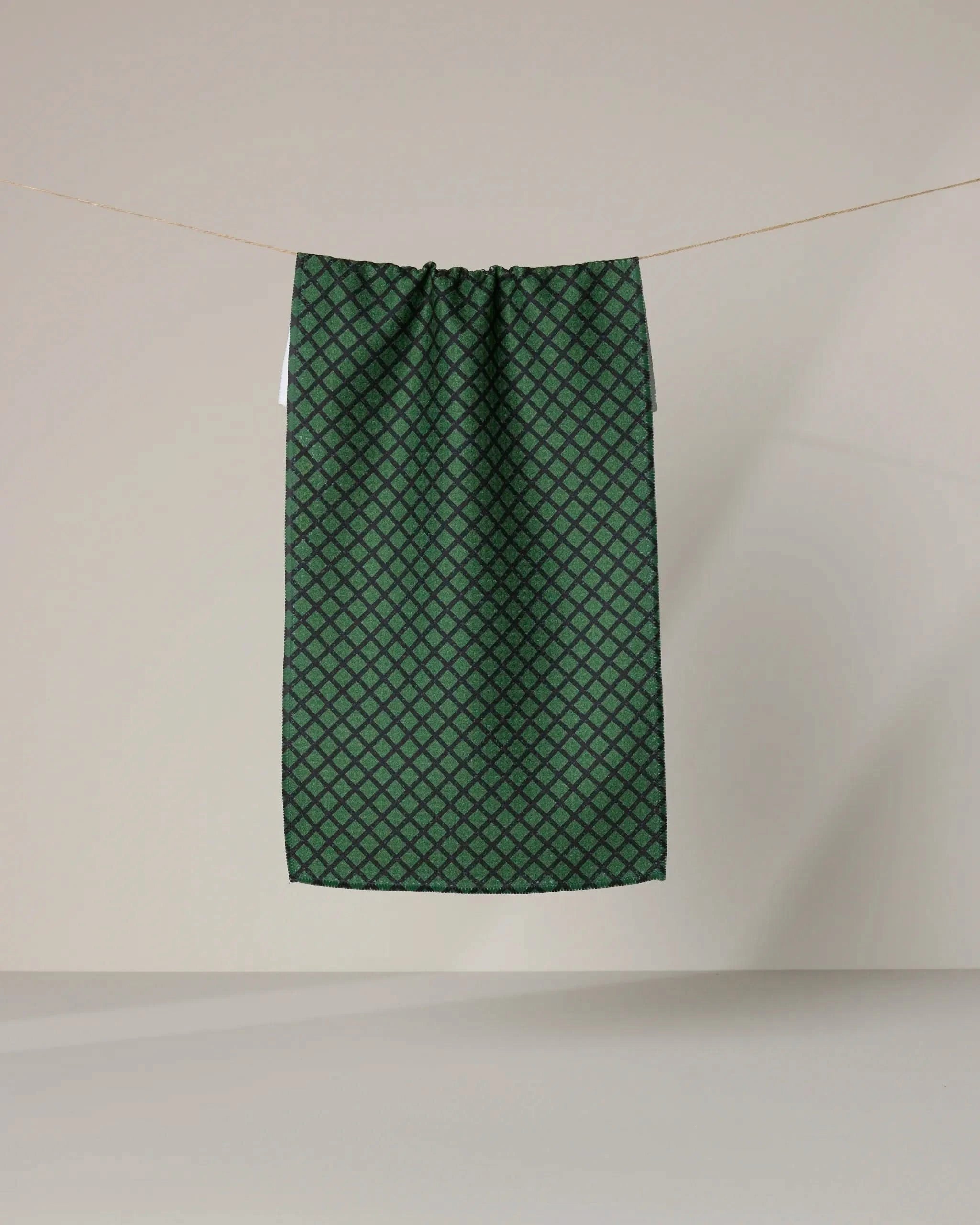 The Geometry Green Lattice towel, a dark green rectangular ultra-absorbent design with a small geometric pattern, hangs on a clothesline against a plain beige background.
