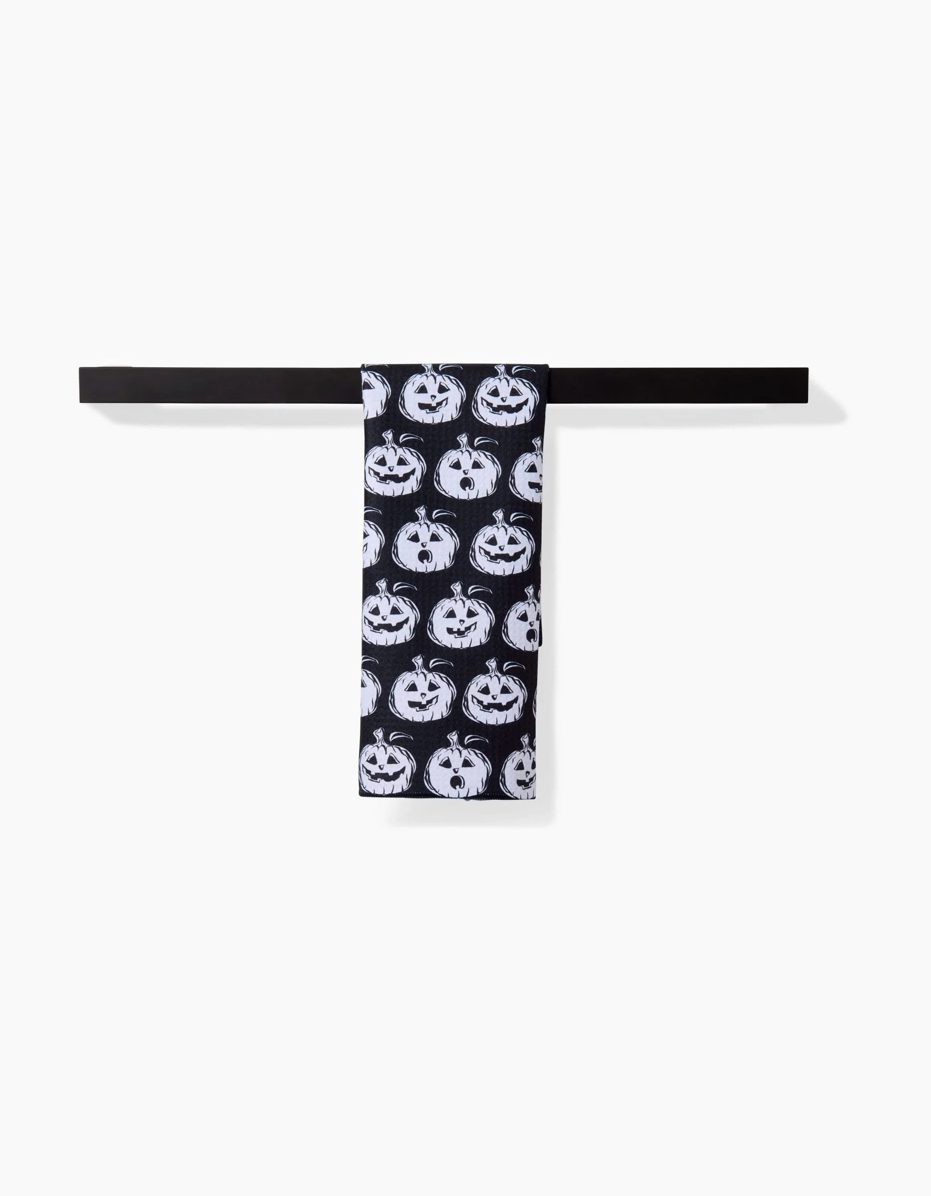 A black bar towel by Aubrey Fairchild hangs on a black bar, displaying the Halloween Pumpkins pattern—white jack-o-lantern faces repeating on a white background.