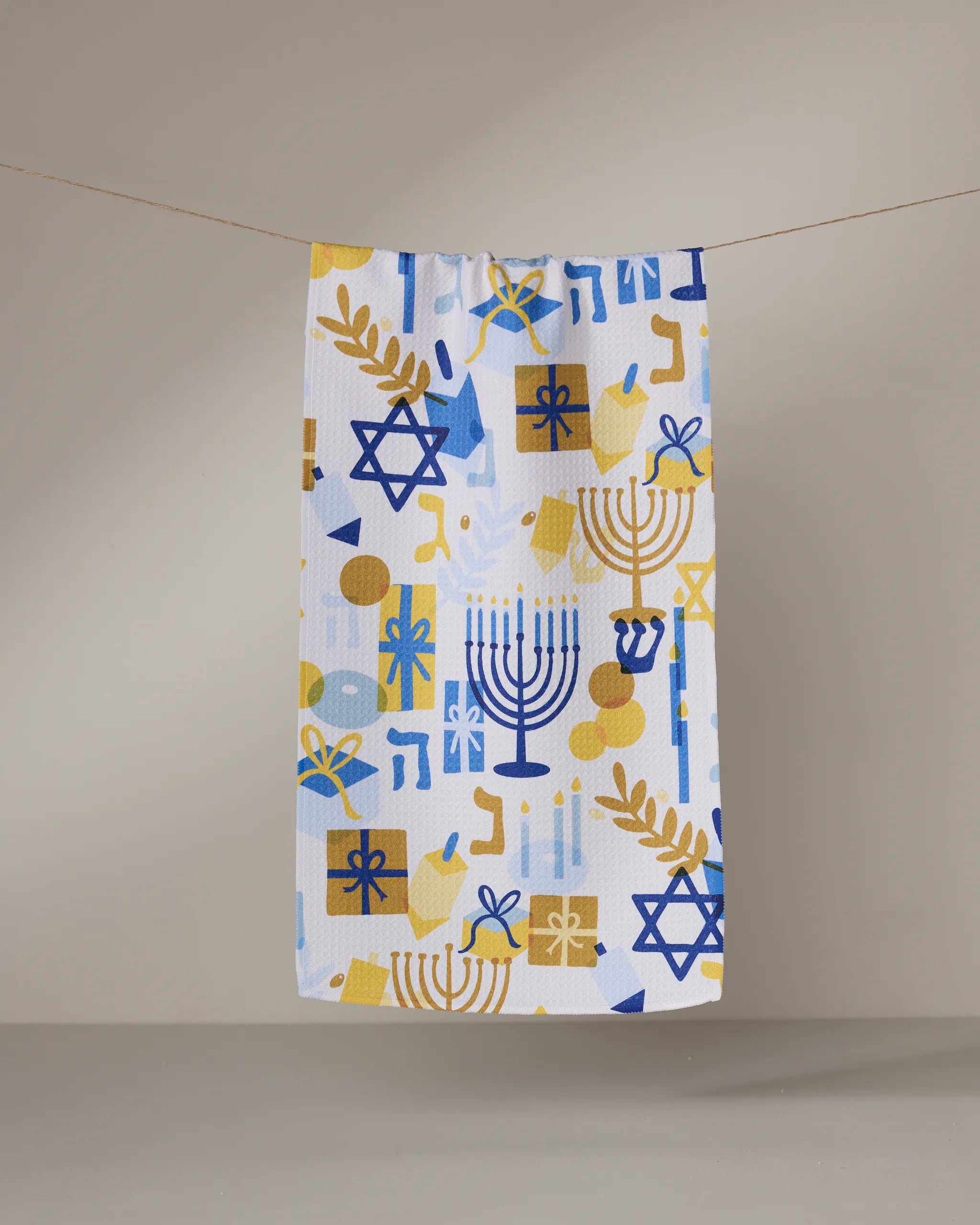 The "Hanukkah is Here" bar towel by Geometry features ultra-absorbent fabric and a festive Hanukkah-themed design with blue and gold menorahs, Stars of David, dreidels, and more, displayed against a simple background.