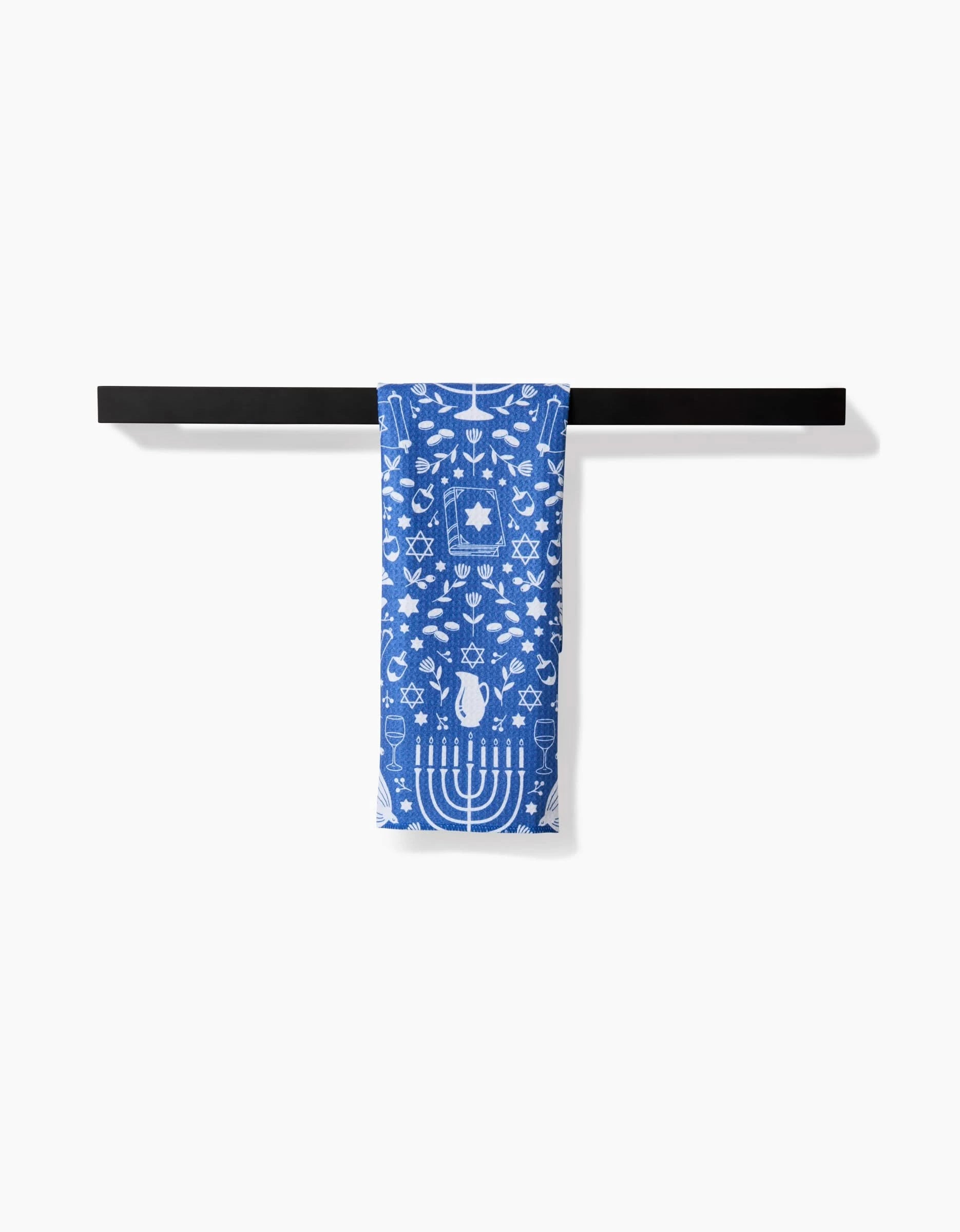 The Hanukkah Sameach kitchen towel by Megan Kampa, featuring blue ultra-absorbent fabric with white menorahs, dreidels, and Stars of David, hangs on a black rack against a white background.