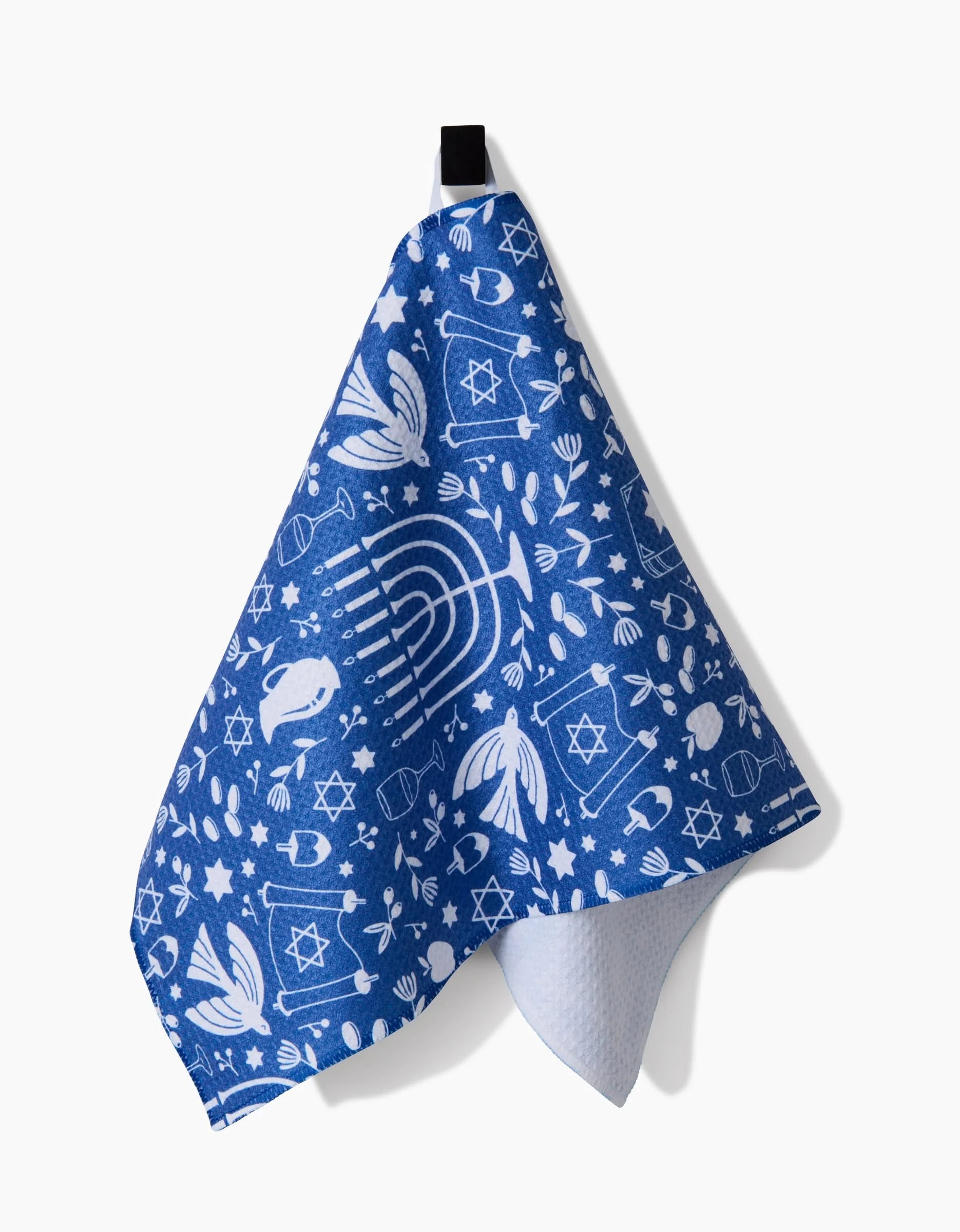 The Hanukkah Sameach towel by Megan Kampa, featuring white Hanukkah-themed designs on blue fabric, hangs from a black loop against a white background.