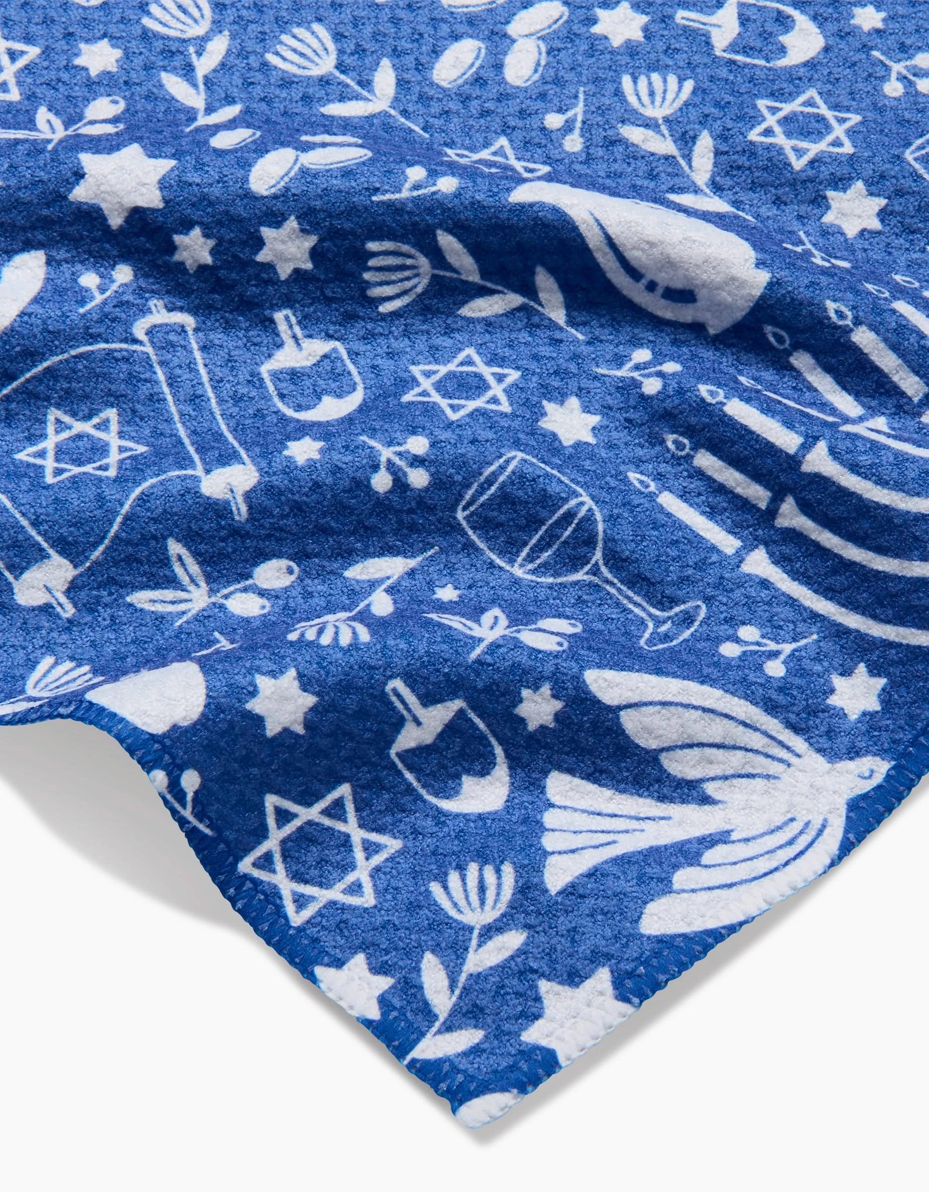 Hanukkah Sameach by Megan Kampa features a blue fabric with white Hanukkah icons—menorahs, dreidels, wine glasses, Stars of David—scattered in a festive pattern. Great for use as a recycled kitchen towel or ultra-absorbent bar towel.