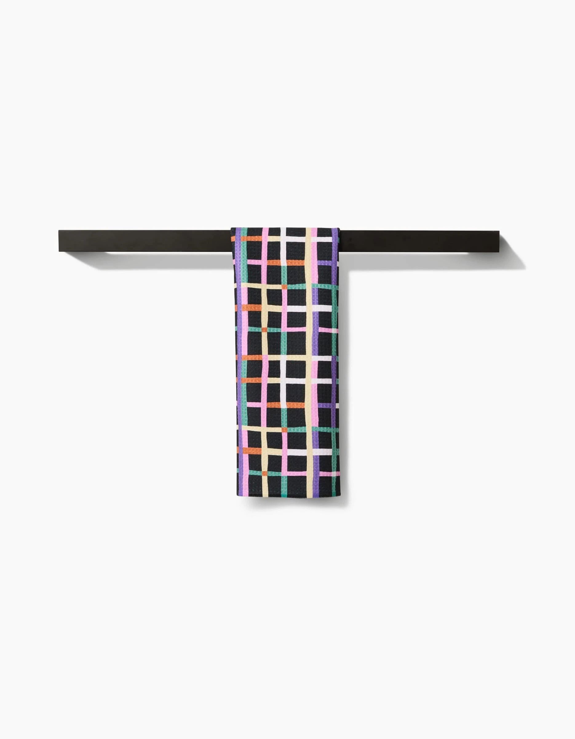 The Happy Plaid bar towel by Claire Iglesias, made from soft Geoweave fabric and featuring a colorful geometric grid design, hangs neatly over a minimalist black towel bar against a plain white background.