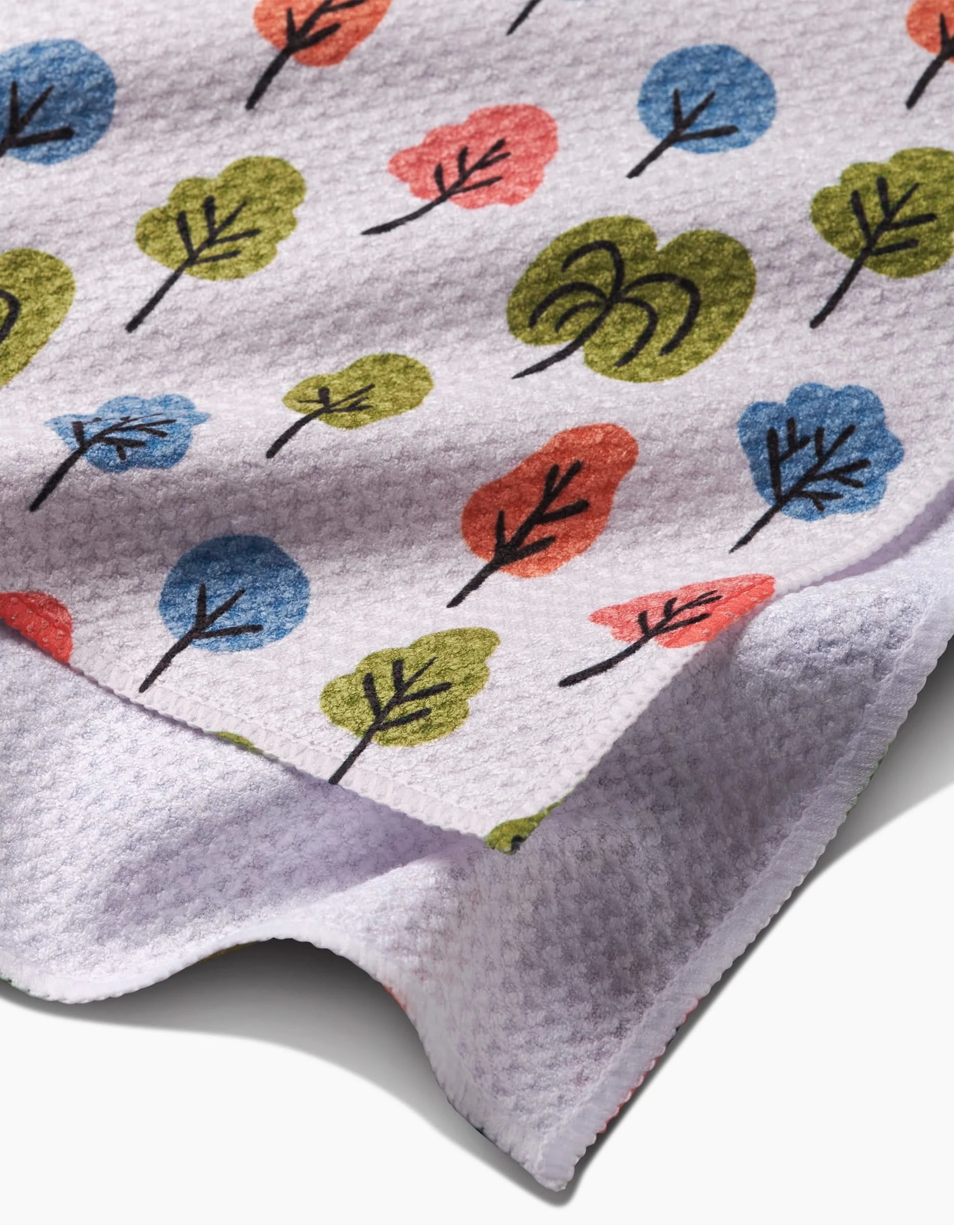 A close-up of the Kayla Ann Happy Trees Bar Towel in textured white Geoweave fabric, showing stylized blue, green, and red trees with black branches and an abstract green shape; the fabrics edge is slightly curled.