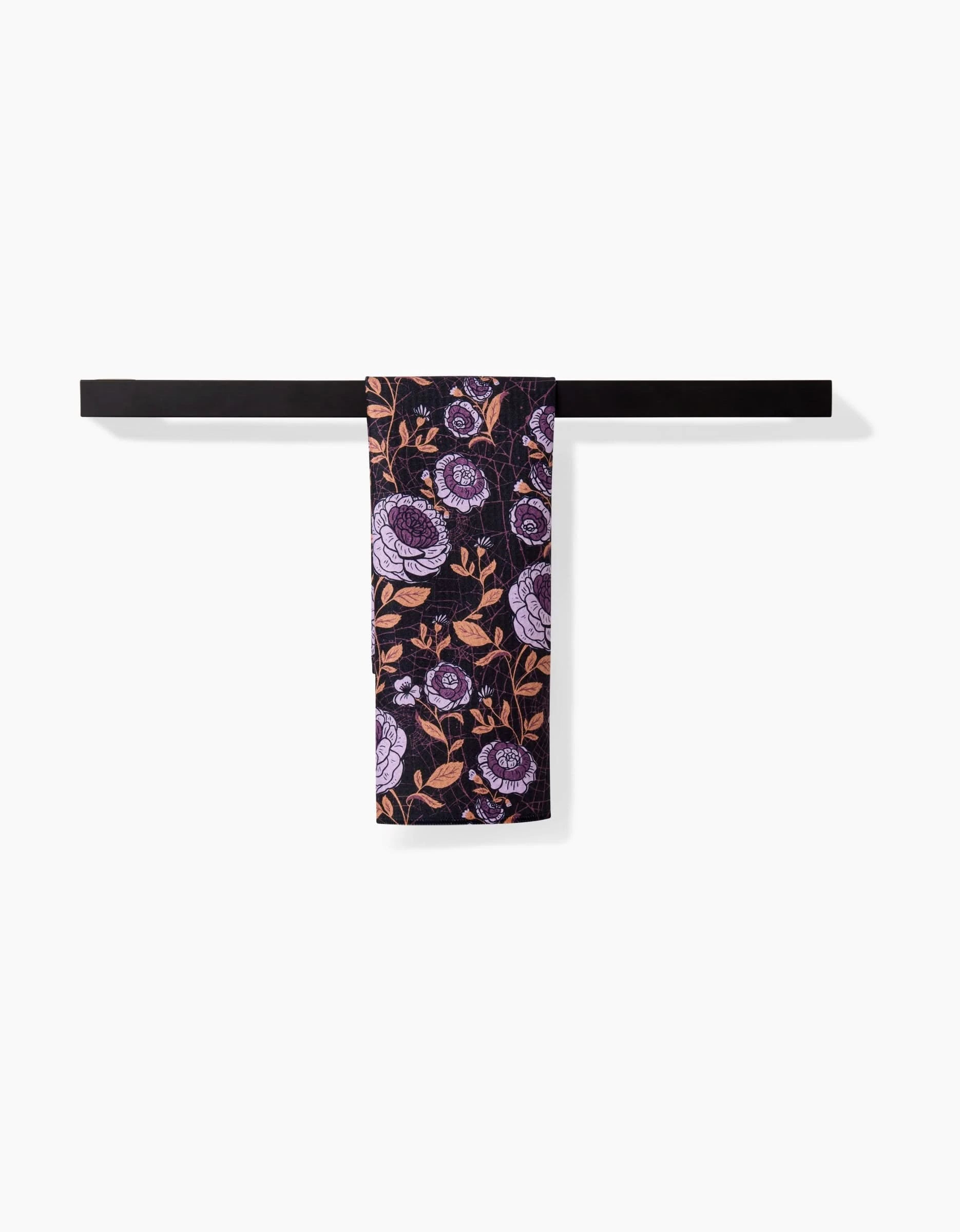 A black towel bar holds a Danelys Sidron Haunted Roses ultra-absorbent towel, featuring a floral pattern with purple and pink flowers and leaves on a dark background, set against a white wall.