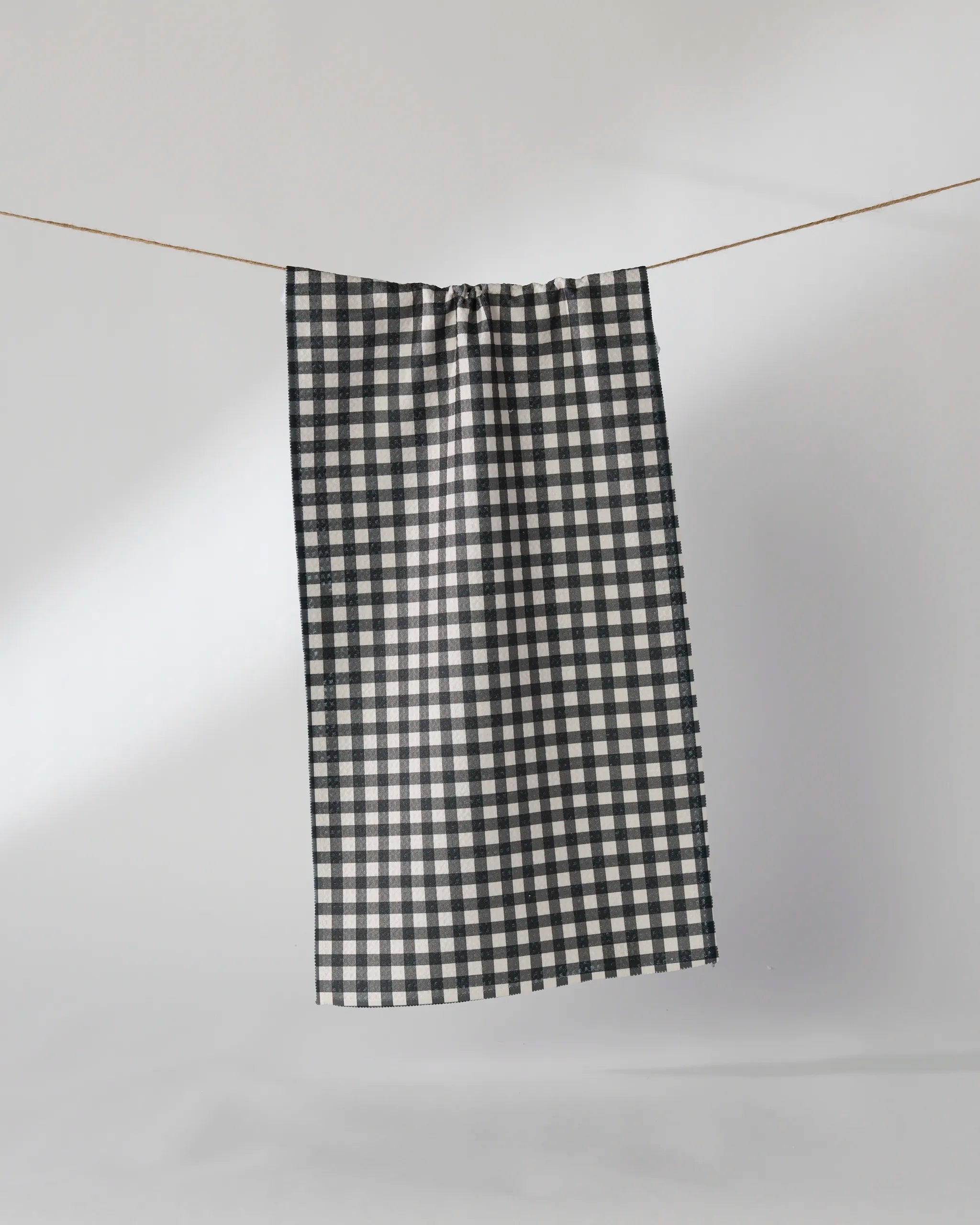 The Geometry Heritage recycled kitchen towel, featuring a black and white checkered design, hangs from a thin string against a plain, light gray background.