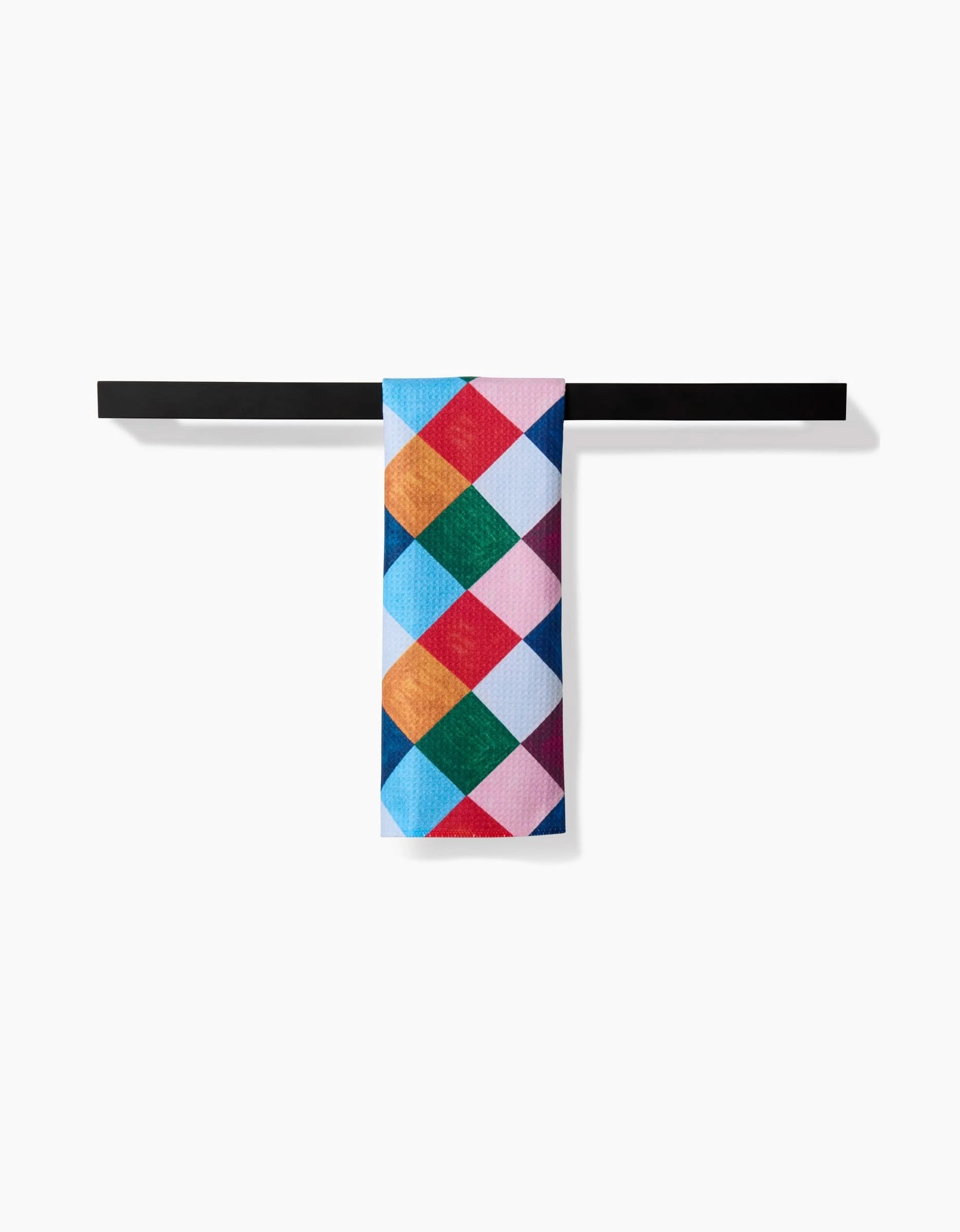 The Holiday Harlequin towel by Daria Solak, featuring a vibrant geometric diamond pattern, is draped over a sleek black bar against a plain white background.