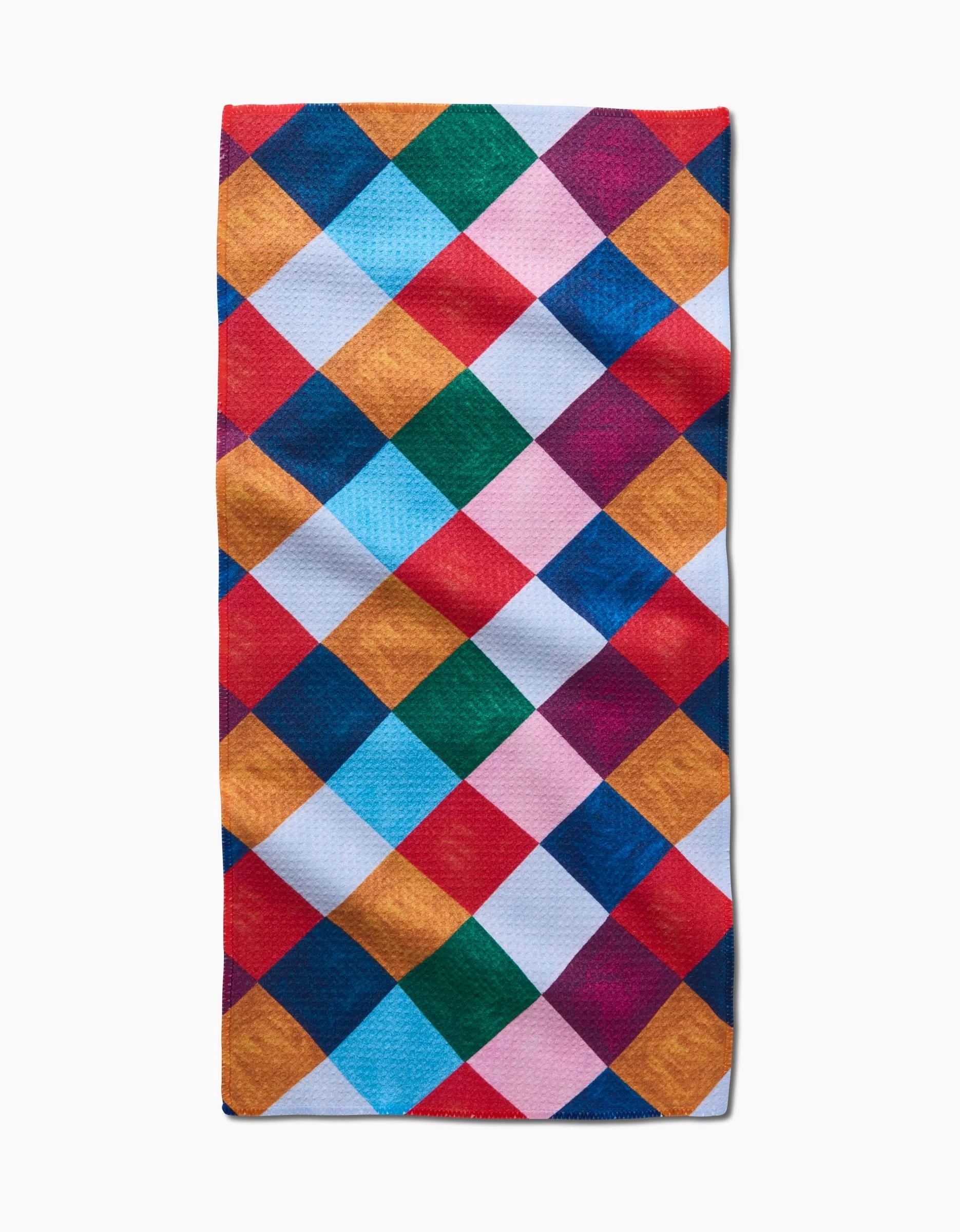 The Holiday Harlequin towel by Daria Solak is a rectangular, ultra-absorbent towel featuring a colorful diamond pattern in red, blue, orange, pink, white, and green on a plain white background.