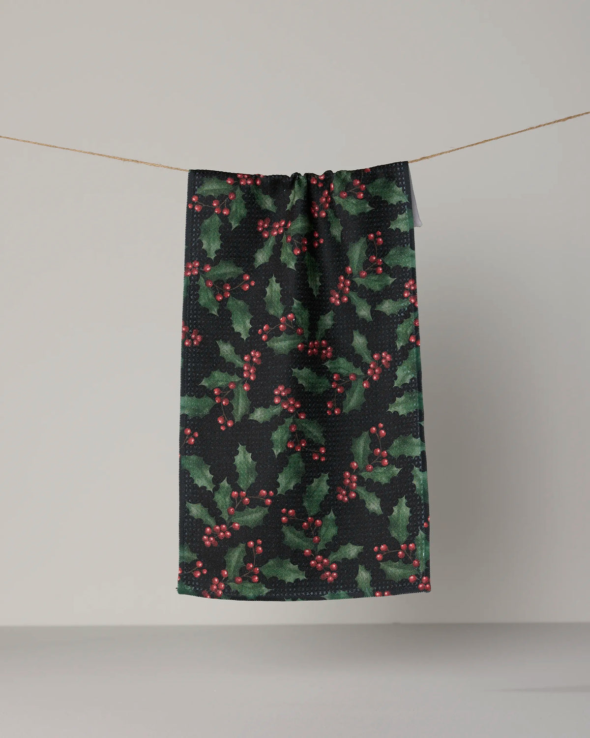 A “Holly Night” mini towel from Geometry, featuring a festive holly and red berry pattern on soft Geoweave fabric, hangs on a string against a plain background.