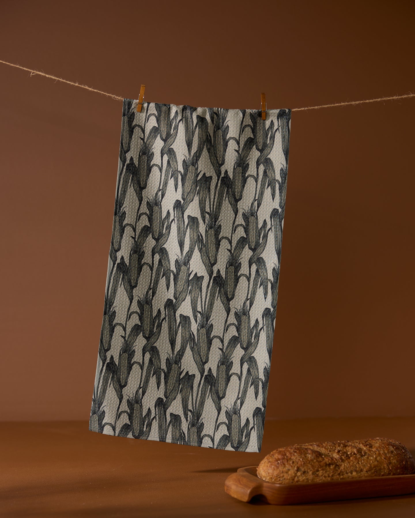 The Geometry Husk bar towel, featuring a black and gray leaf pattern and made from recycled materials, hangs on a clothesline with clothespins. In front, a loaf of bread sits on a wooden board against a brown backdrop.