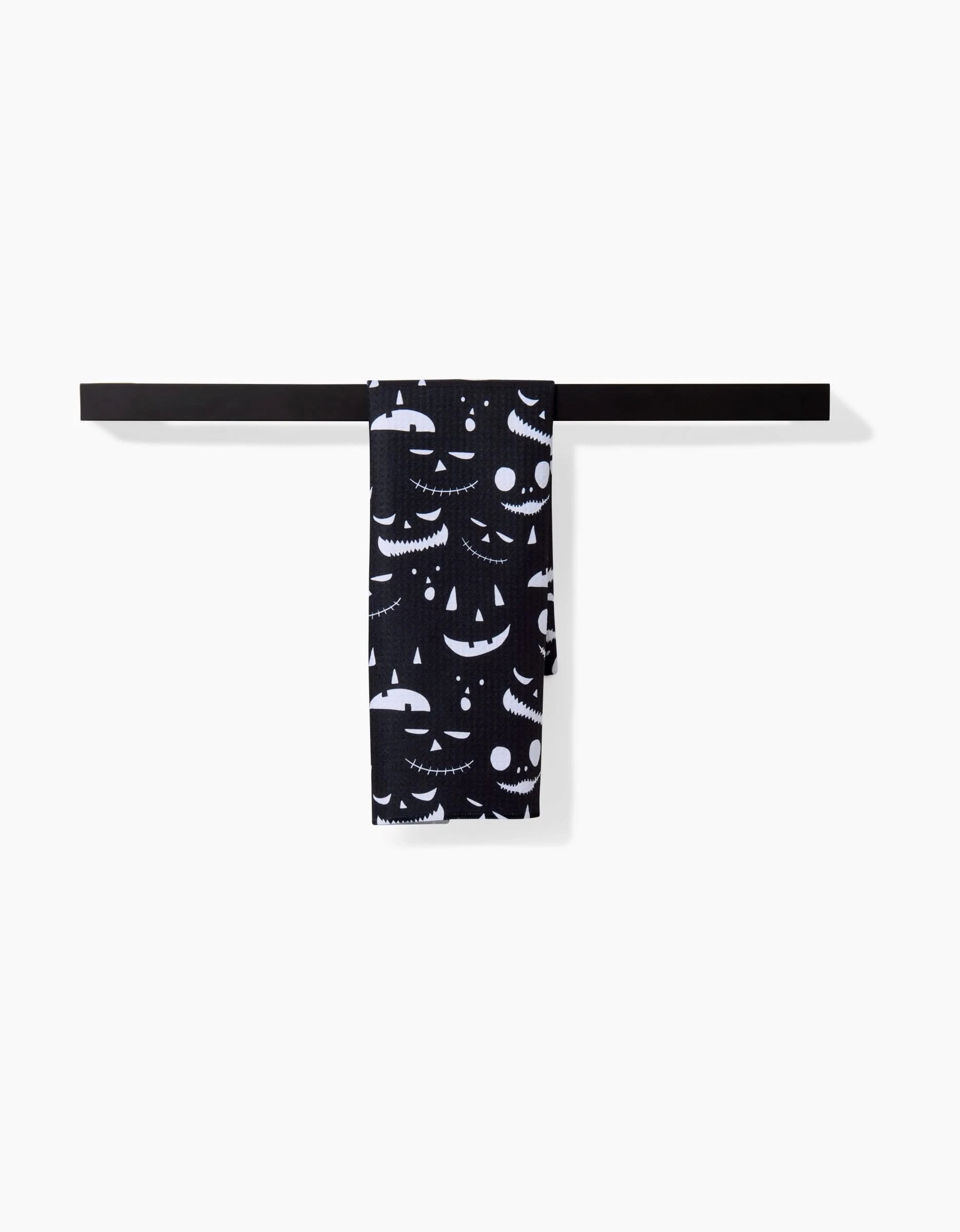 The Danelys Sidron Jack O Lantern Jamboree Black towel, with white pumpkin faces and made from ultra-absorbent recycled materials, hangs neatly over a minimalist black rack against a plain white background.