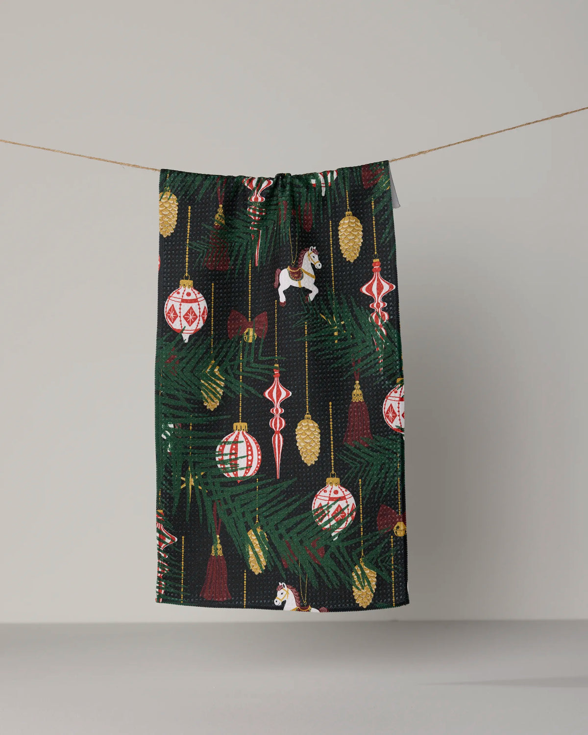 The Geometry Keepsake Ornaments mini towel features a festive pattern of ornaments, pine branches, and toy horses in vibrant red, gold, and green. Made from recycled materials, this eco-friendly towel brings holiday cheer and sustainable charm to any space.