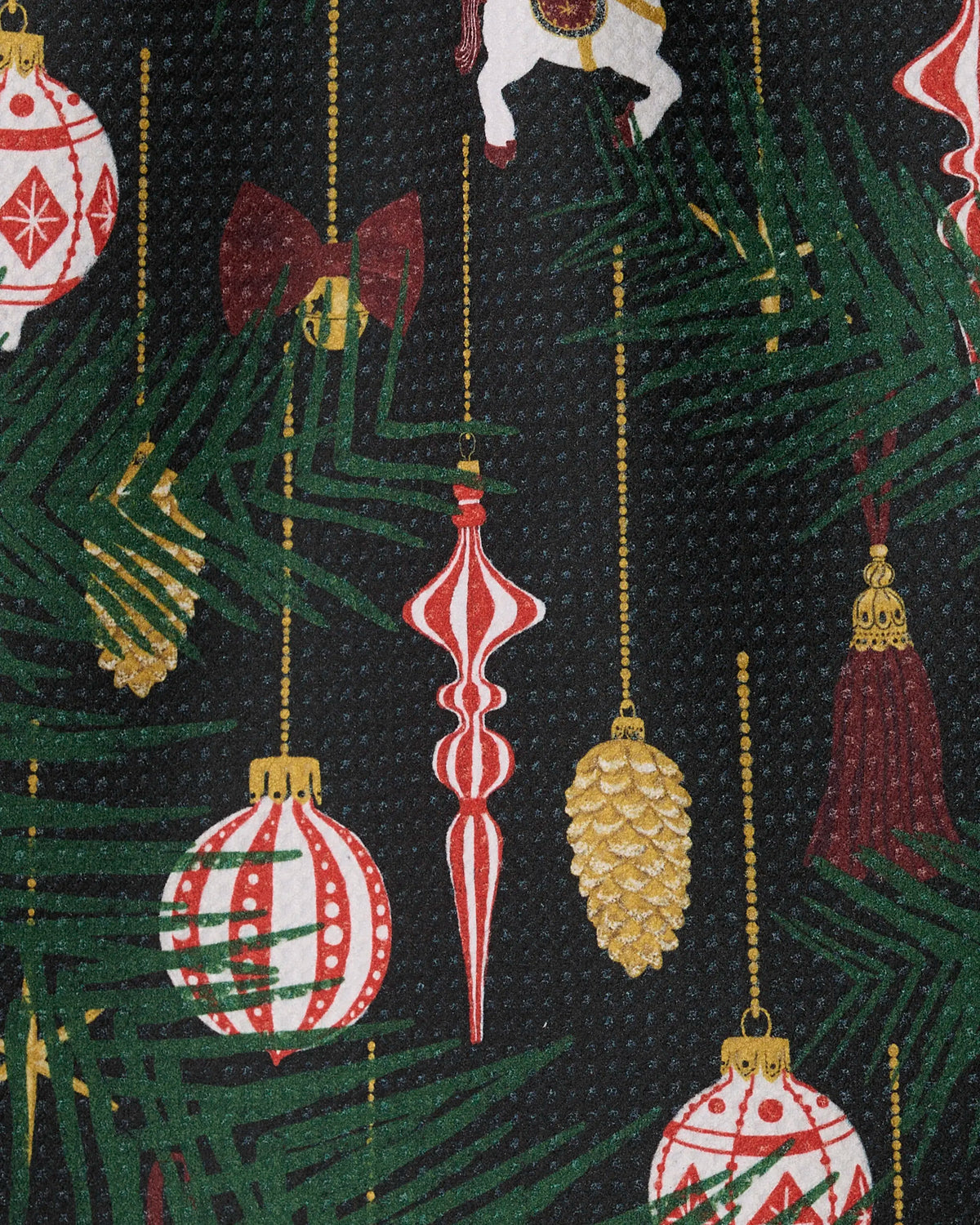 Geometry’s Keepsake Ornaments mini towel features patterned Geoweave fabric adorned with green pine branches, red and gold ornaments, pinecones, bows, a white carousel horse, and tassels against a dark background for a festive touch.