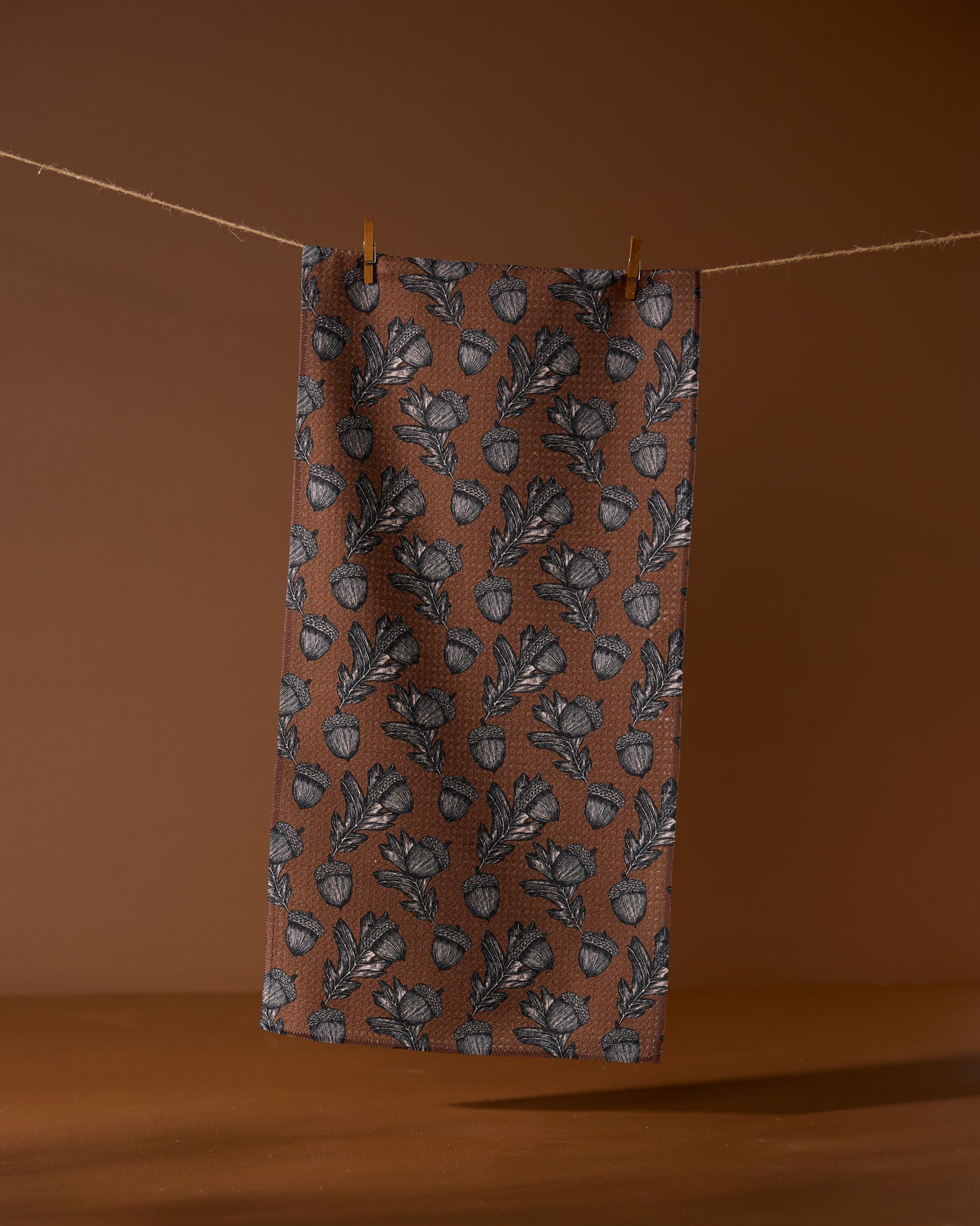 The Geometry Kindred towel, made from recycled materials, features a brown cloth with a repeating black leaf and acorn pattern. It hangs from a string with two clothespins against a brown background.