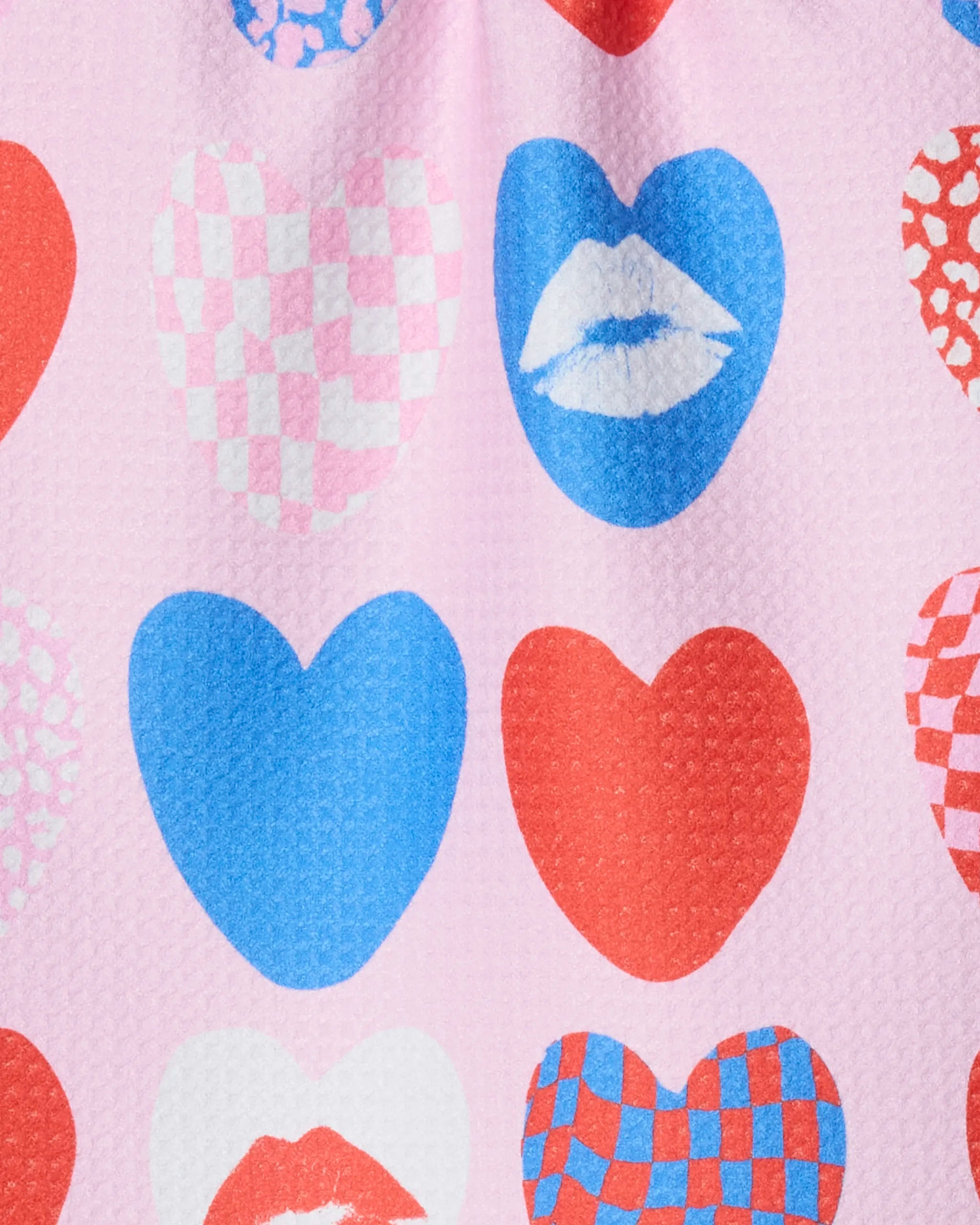 The Kiss and Tell fabric by Geometry features a playful, pink recycled material printed with blue and red hearts—some with checkerboards or lips—in a whimsical, repeating design.