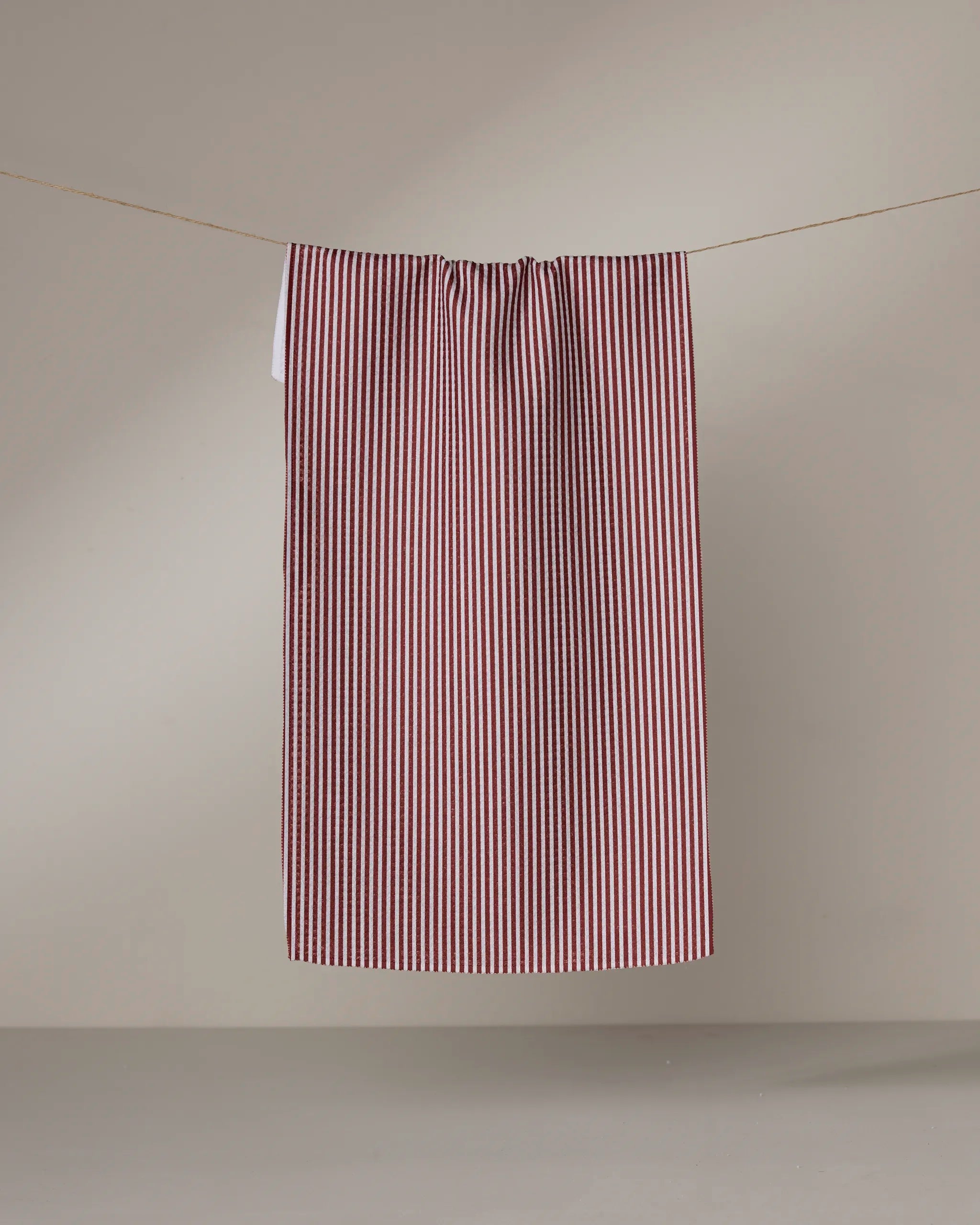 The Geometry Kringle bar towel, featuring red and white vertical stripes and made from recycled materials, hangs on a clothesline against a plain light gray background.