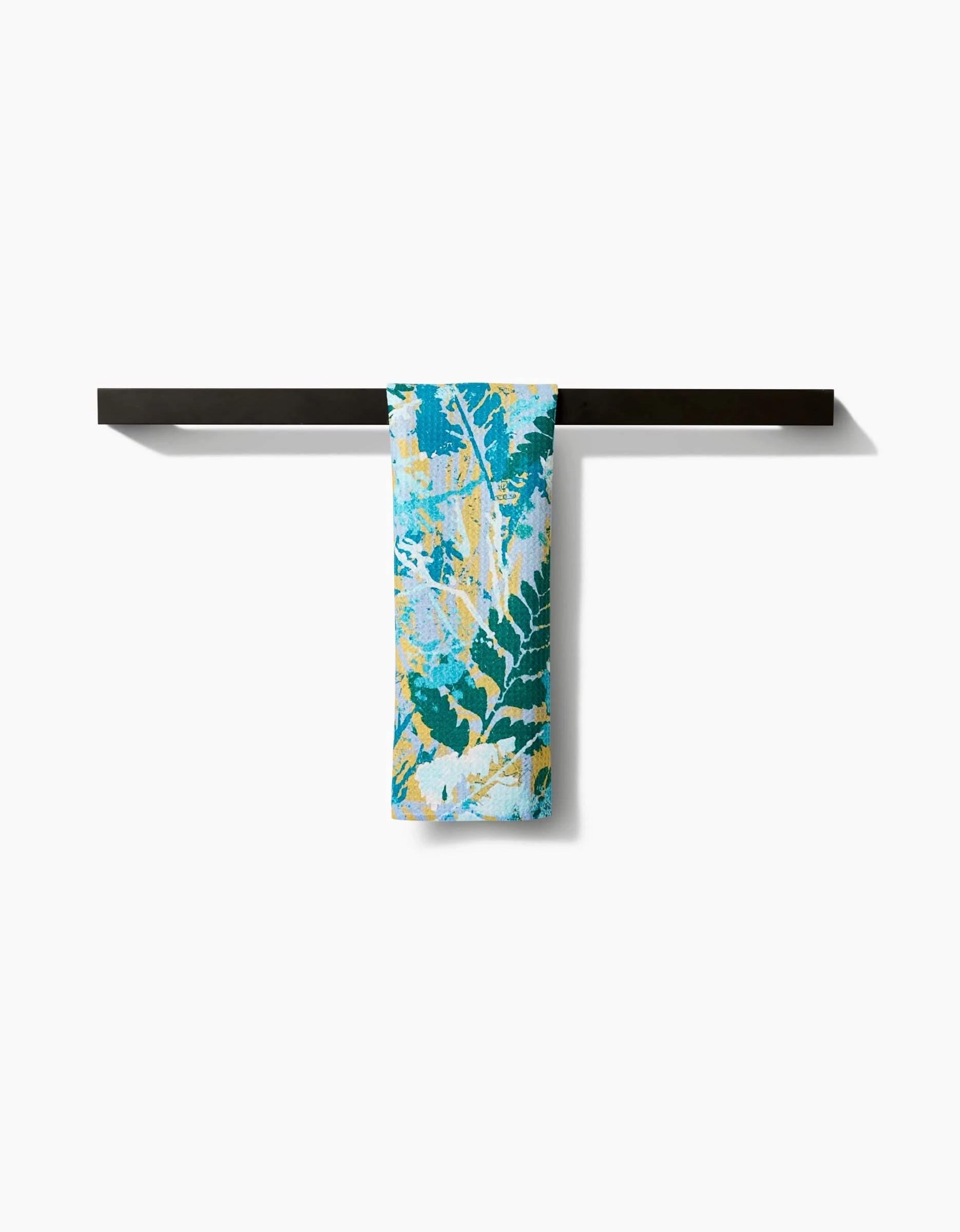 The Leafy Bush bar towel by Taryn Brennan features a vibrant blue, green, and yellow leaf pattern on Geoweave fabric, hanging neatly on a dark towel bar and standing out against a crisp white background.