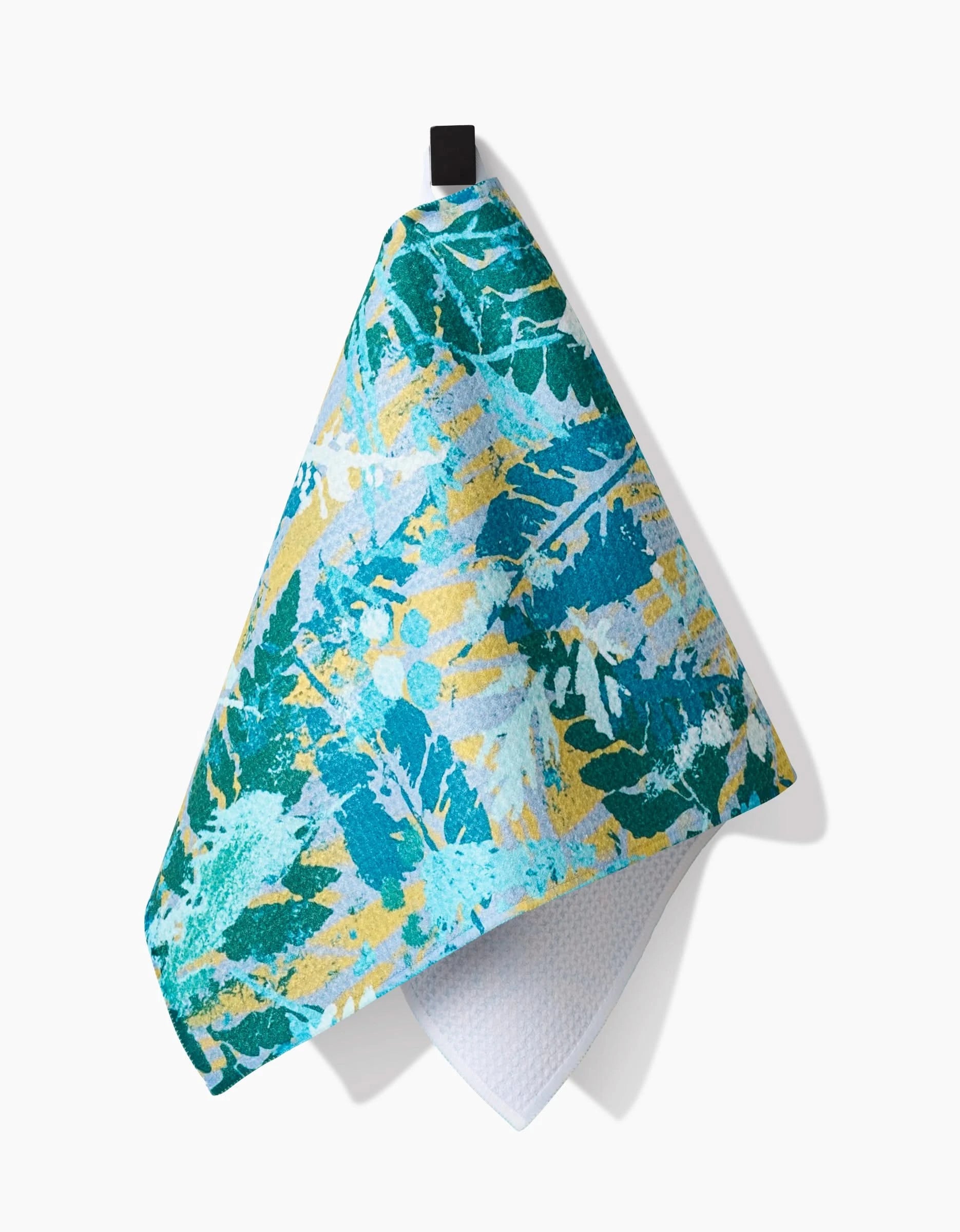 The Taryn Brennan Leafy Bush Bar Towel in blue, green, and yellow leaf patterns hangs from a black loop. Its Geoweave fabric front is vibrant; the textured white underside shows against a white background.
