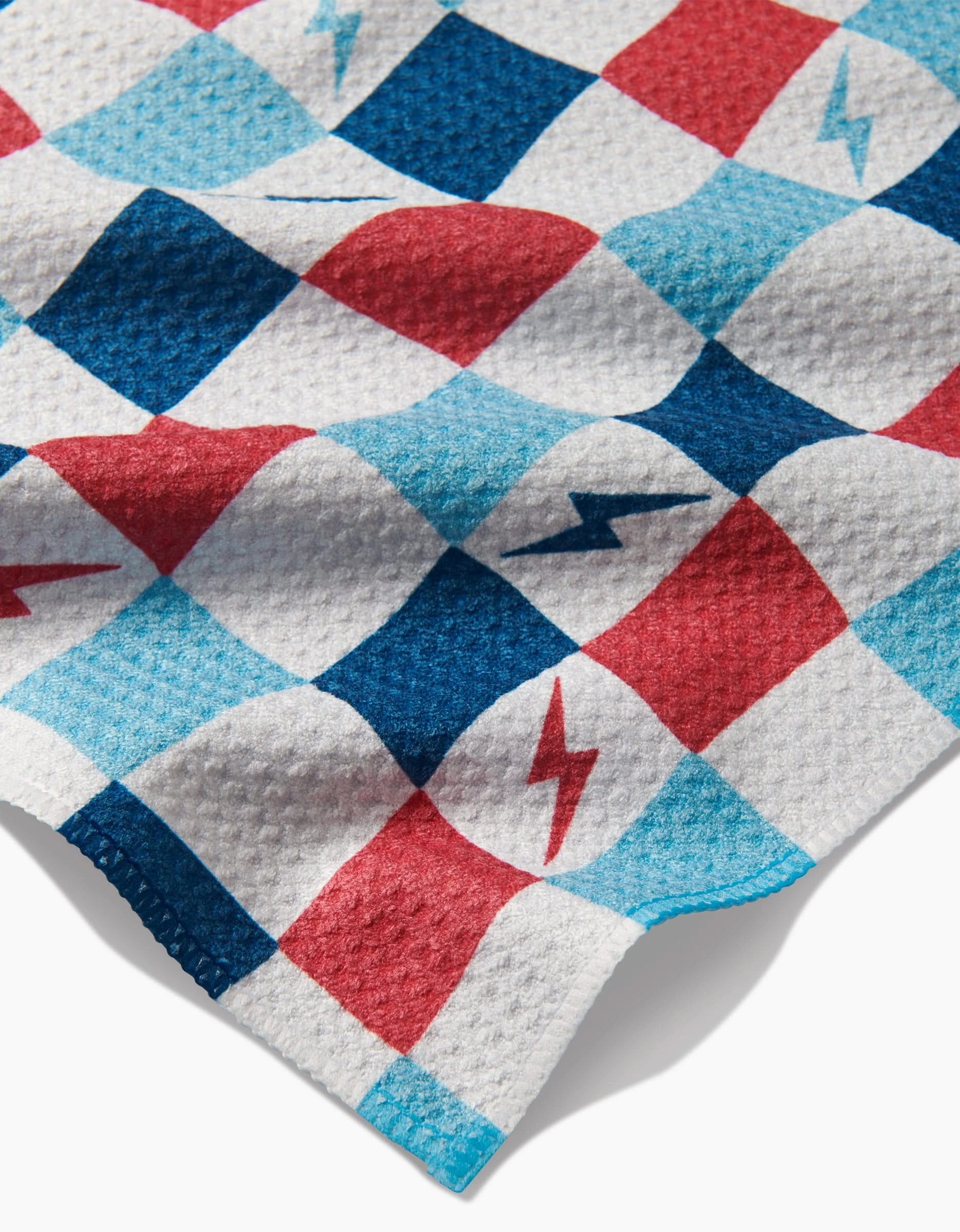 A close-up of Hufton Studios Lightening Bolt Chess bar towel, made from Geoweave fabric, featuring a blue, red, and white checkered pattern with some squares displaying blue or red lightning bolt symbols. The soft material appears slightly wavy.