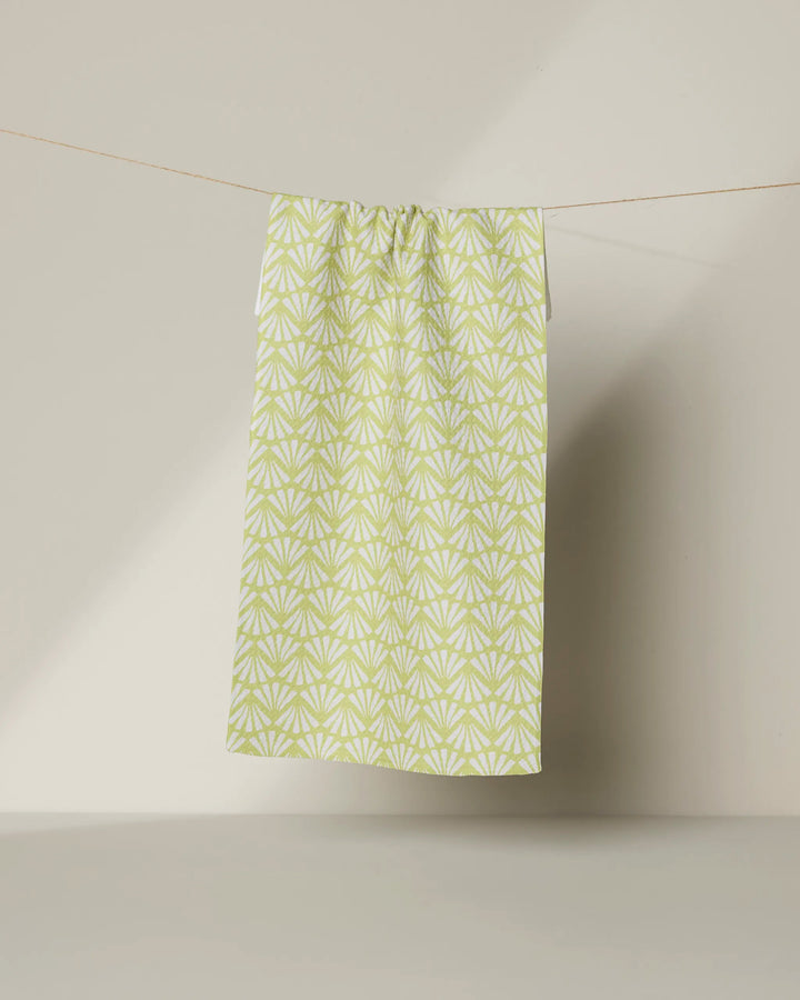 The Geometry Lime Blossom kitchen towel, featuring a green and white repeating geometric fan-like pattern, hangs from a string against a plain, light-colored background.