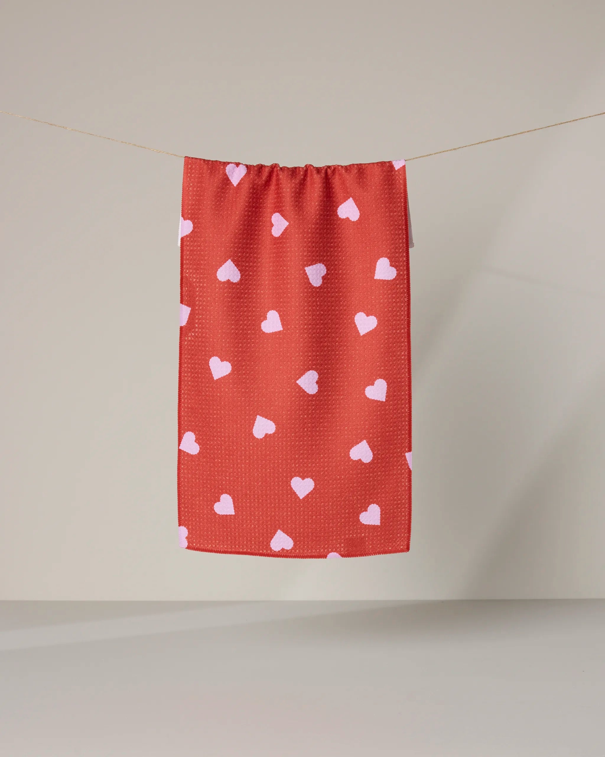 The Geometry Love Toss towel, featuring ultra-absorbent fabric and a red design with white hearts, hangs on a clothesline against a neutral background.