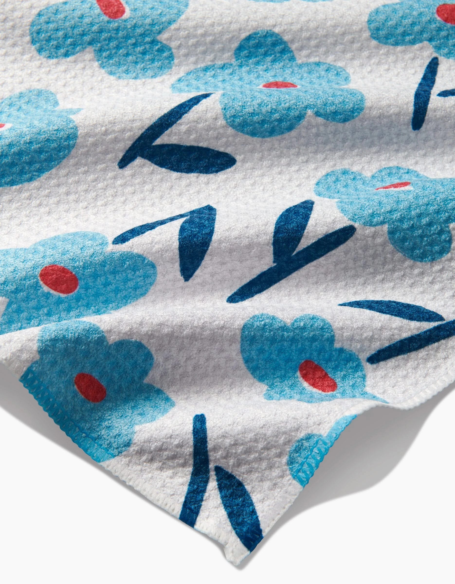 The Luca Daisy bar towel by Hufton Studio features white Geoweave fabric with blue flowers, red centers, and dark blue stems and leaves. One corner of the towel is slightly curled.