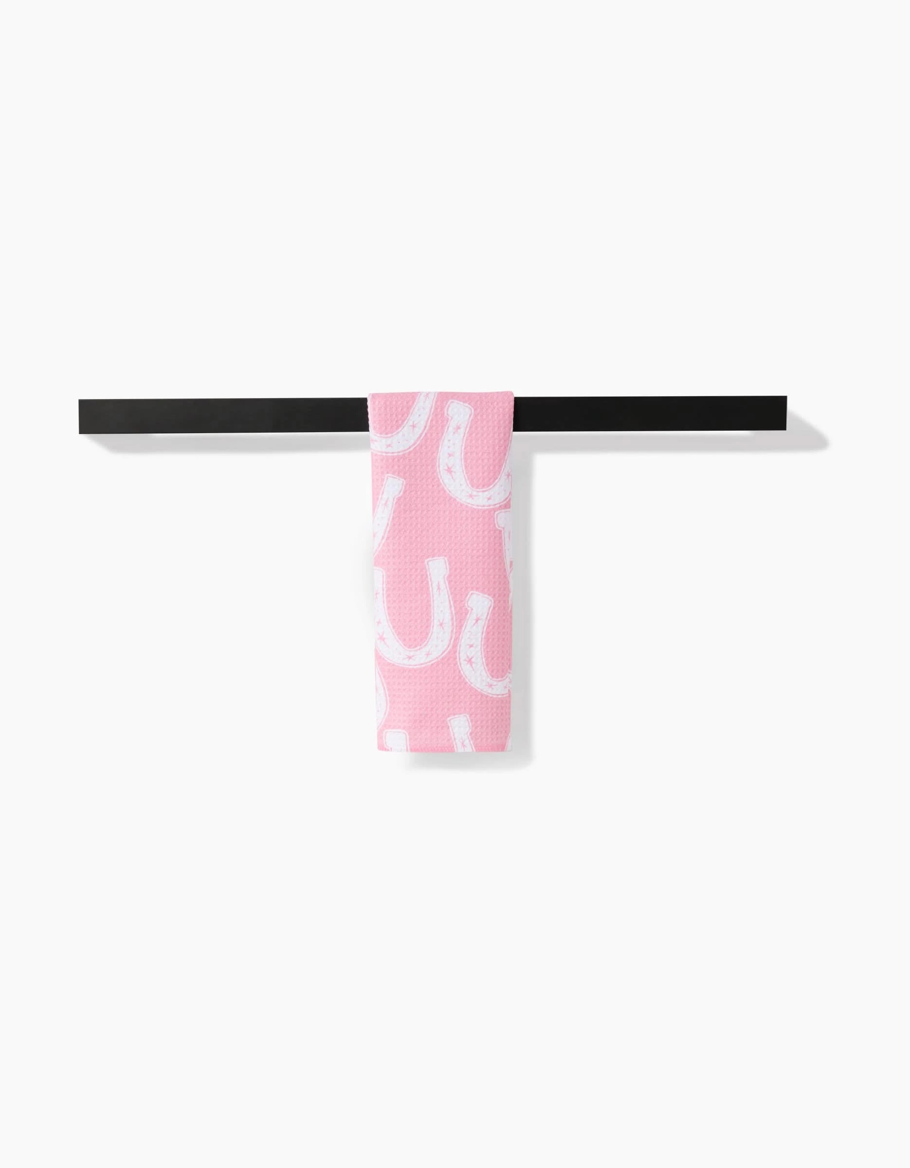 The Lucky Horseshoe bar towel by Aubrey Fairchild, featuring a white horseshoe pattern on pink Geoweave fabric, hangs over a black bar against a plain white background.