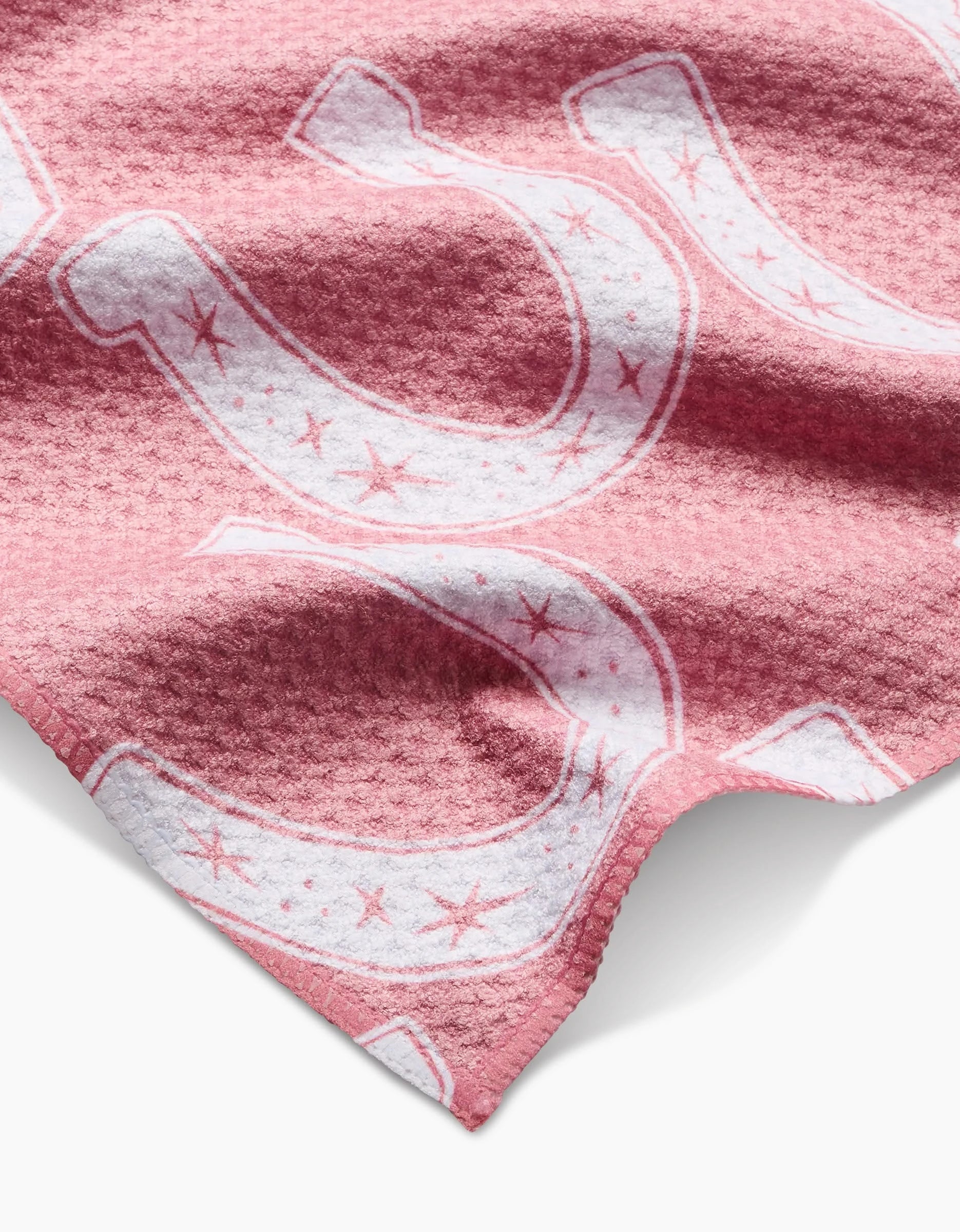 A close-up of the Aubrey Fairchild Lucky Horseshoe bar towel shows its textured pink Geoweave fabric with large white horseshoes and small stars. Made from recycled material, the soft towel has visible folds and shadows.