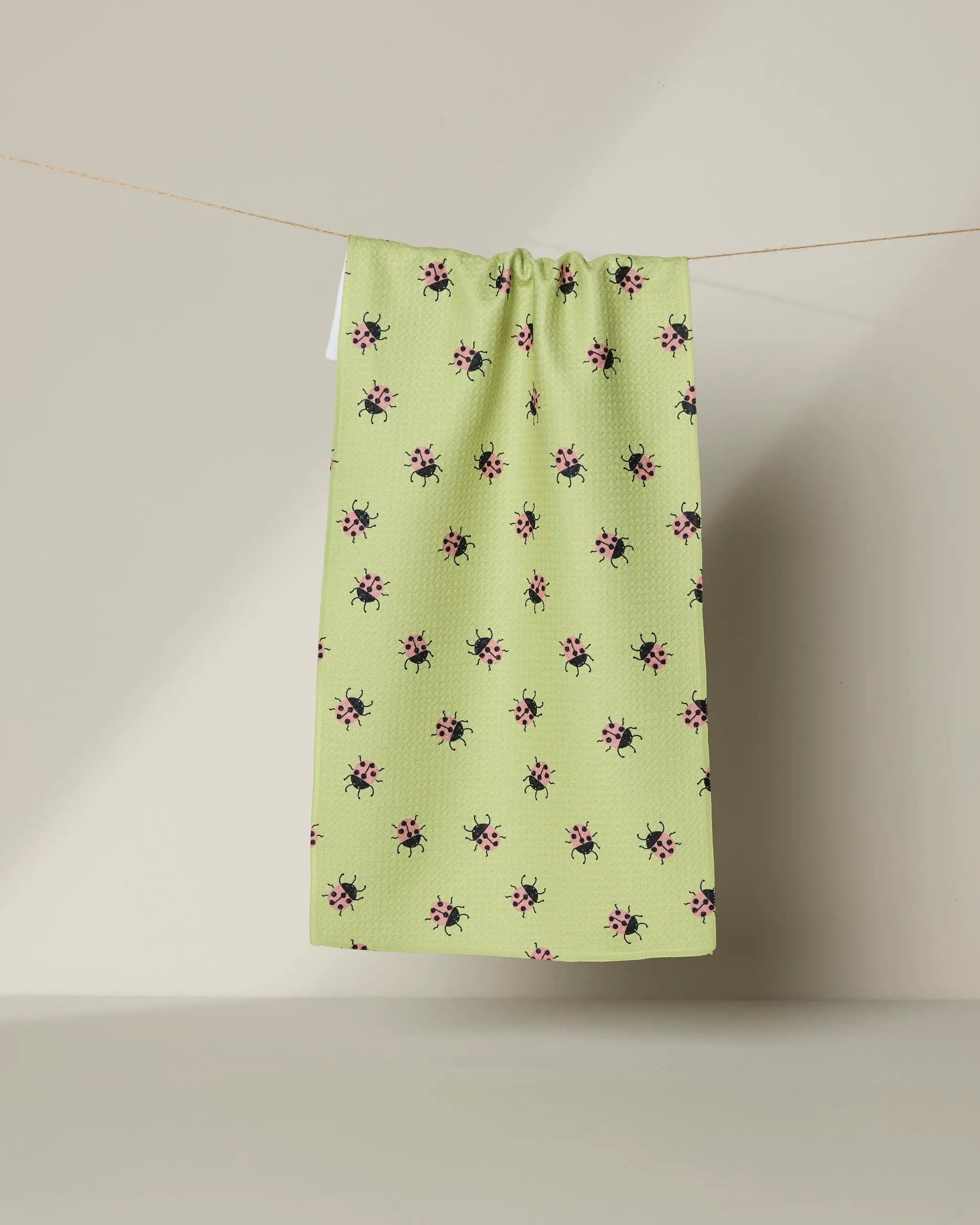 The Mariquita kitchen towel by Geometry features a light green Geoweave fabric with small pink and black floral patterns, shown hanging on a thin string against a neutral background.