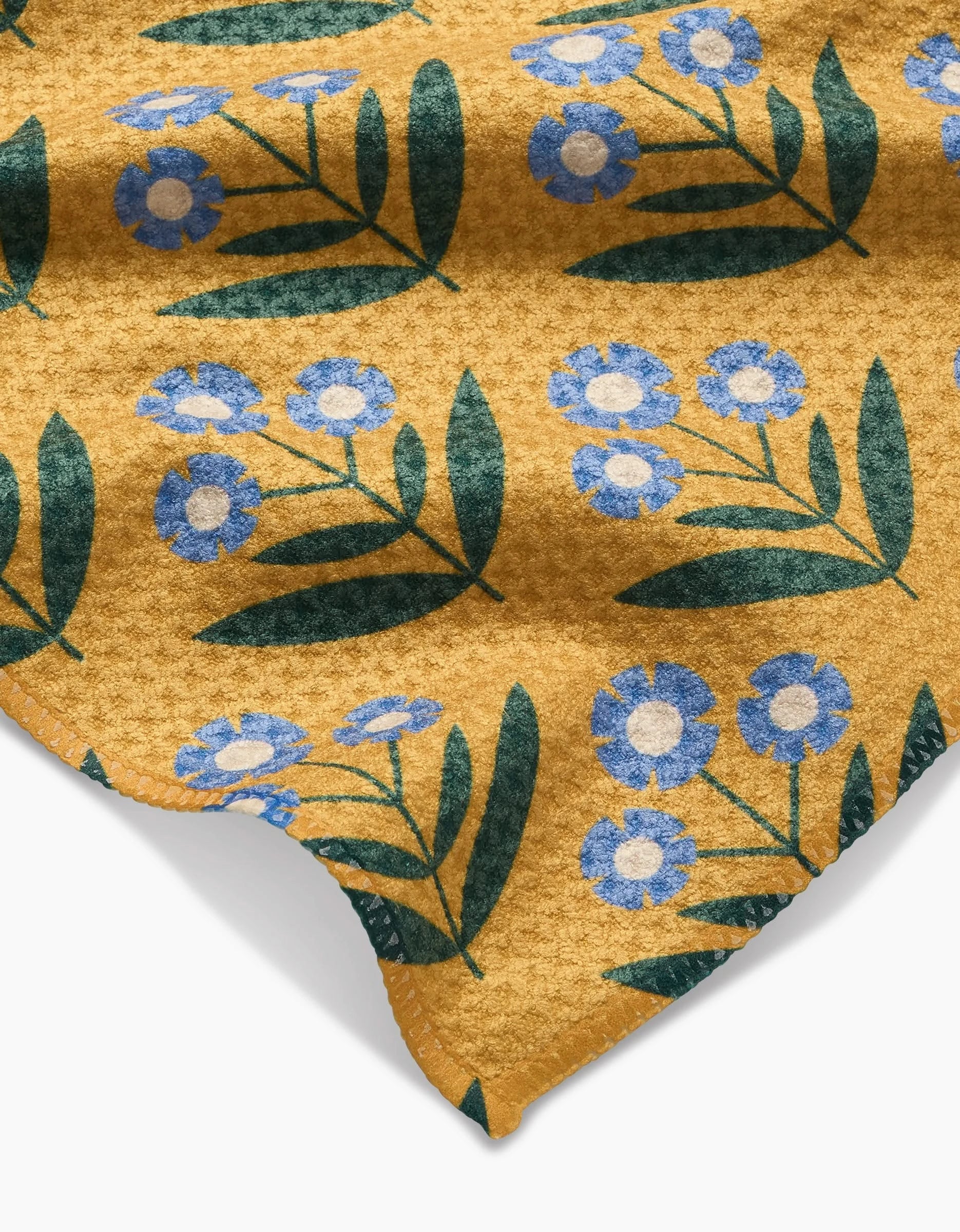 The Mayday by Megan Kampa is a yellow Geoweave fabric with blue flowers, dark green leaves, a subtly textured surface, and a wavy edge in the bottom corner.