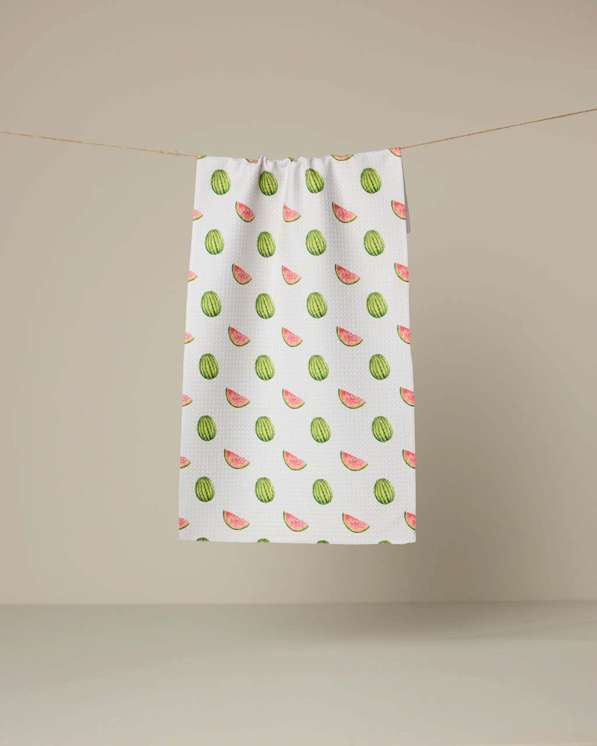 The Geometry Melon Market Scattered bar towel, made from recycled materials and featuring a playful whole watermelon and slice pattern, hangs on a clothesline against a beige background.