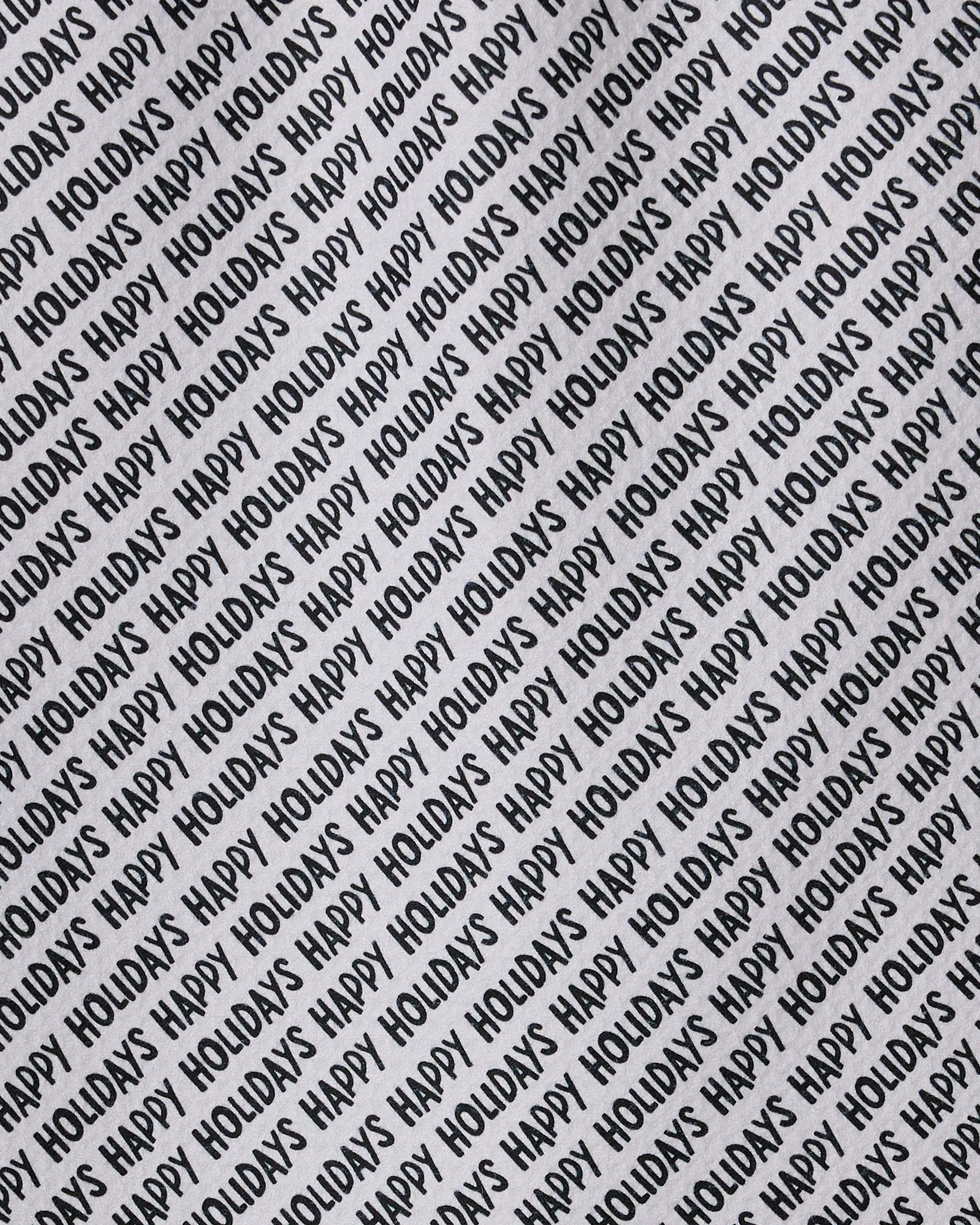 The Merrier towel by Geometry features diagonal black "HAPPY HOLIDAYS" text on a white background, creating a festive, patterned look. Ultra-absorbent and made with recycled materials for an eco-friendly touch.