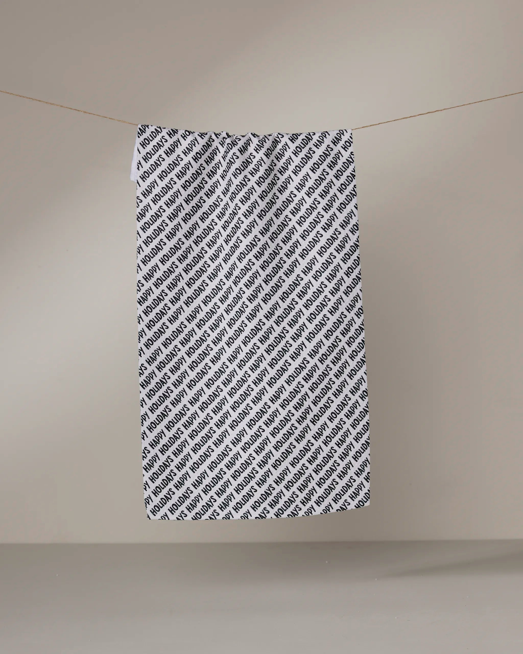 Geometry’s “Merrier” towel, ultra-absorbent and white with “BOSS” printed repeatedly in a diagonal black pattern, hangs from a clothesline against a plain beige background.