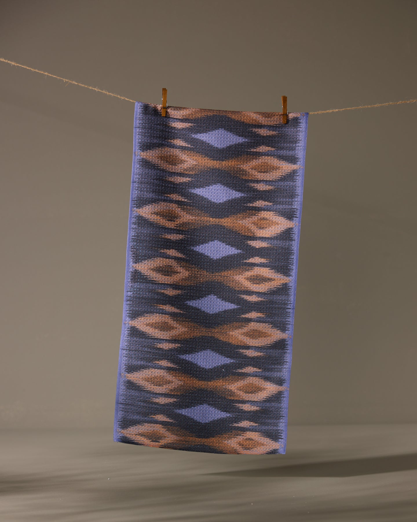 The Geometry Mesa towel, a rectangular ultra-absorbent piece with a blue, brown, and beige geometric pattern, hangs from a clothesline against a plain background, secured by two wooden clothespins.