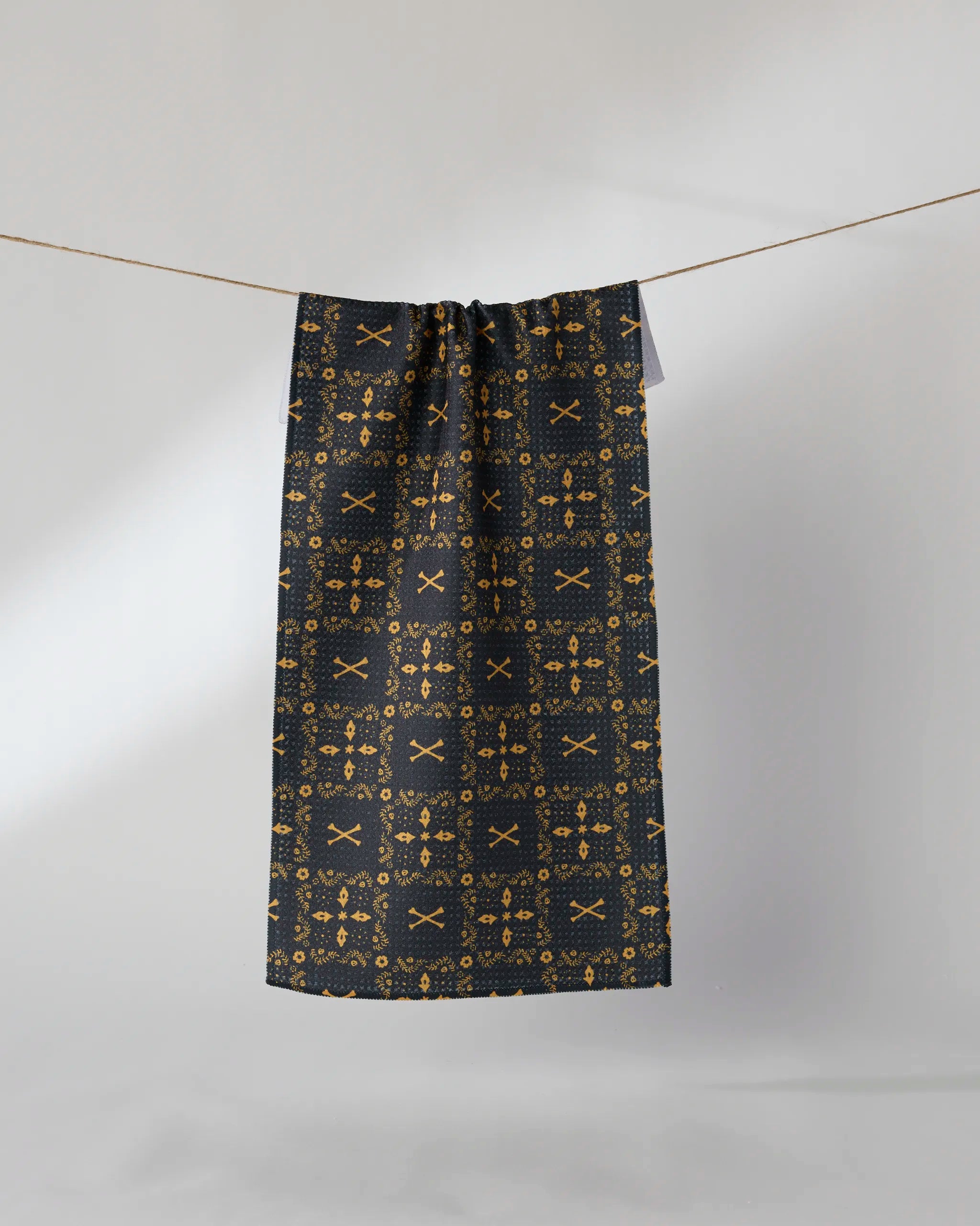 The Geometry Midnight Bandana, an ultra-absorbent dark towel with a gold geometric and floral design, hangs from a clothesline against a plain white background.