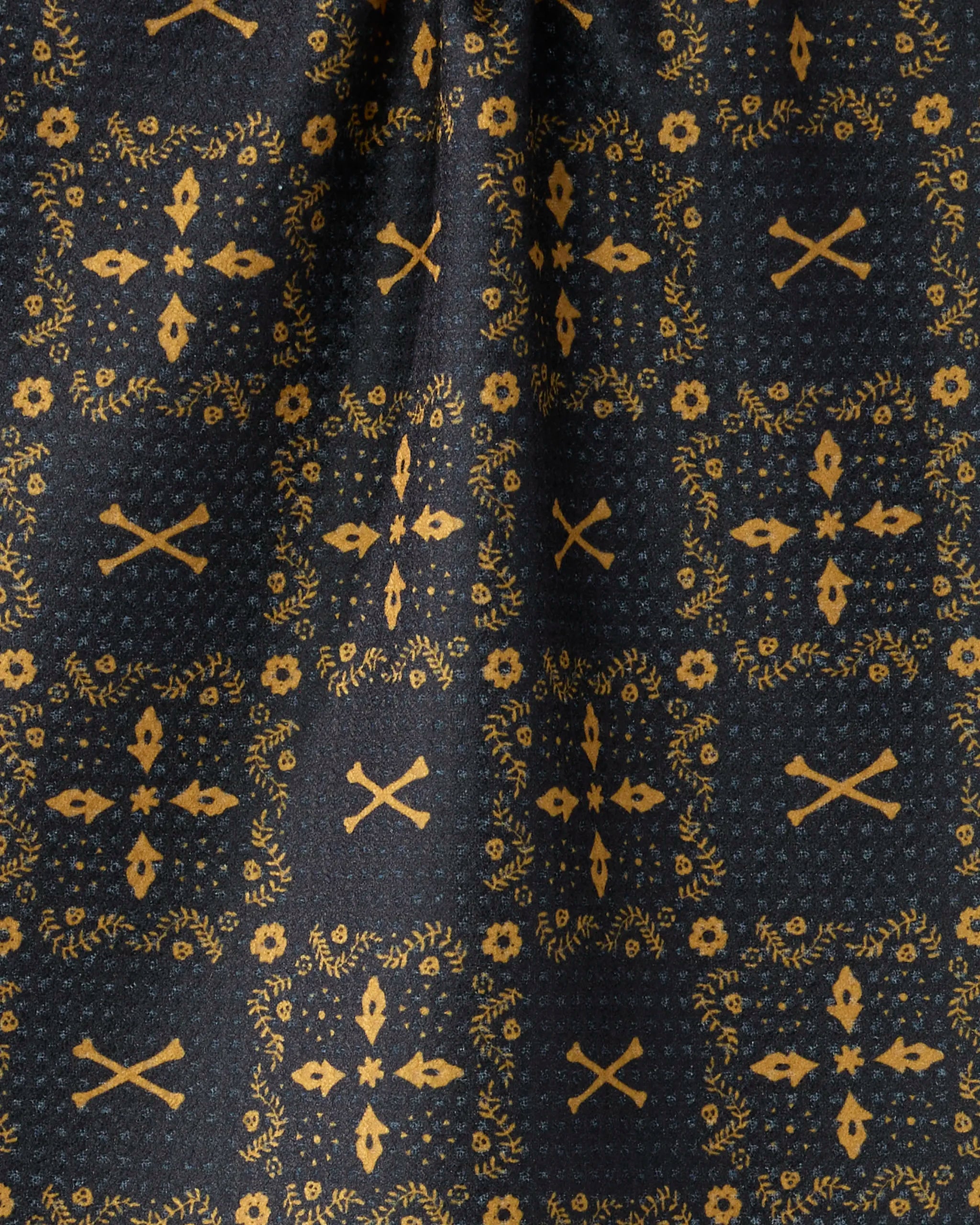 Close-up of the Geometry Midnight Bandana's dark fabric, featuring a repeating gold pattern of crossed rifles, laurel wreaths, and floral motifs in symmetrical squares. A vertical shadow hints at a fold.