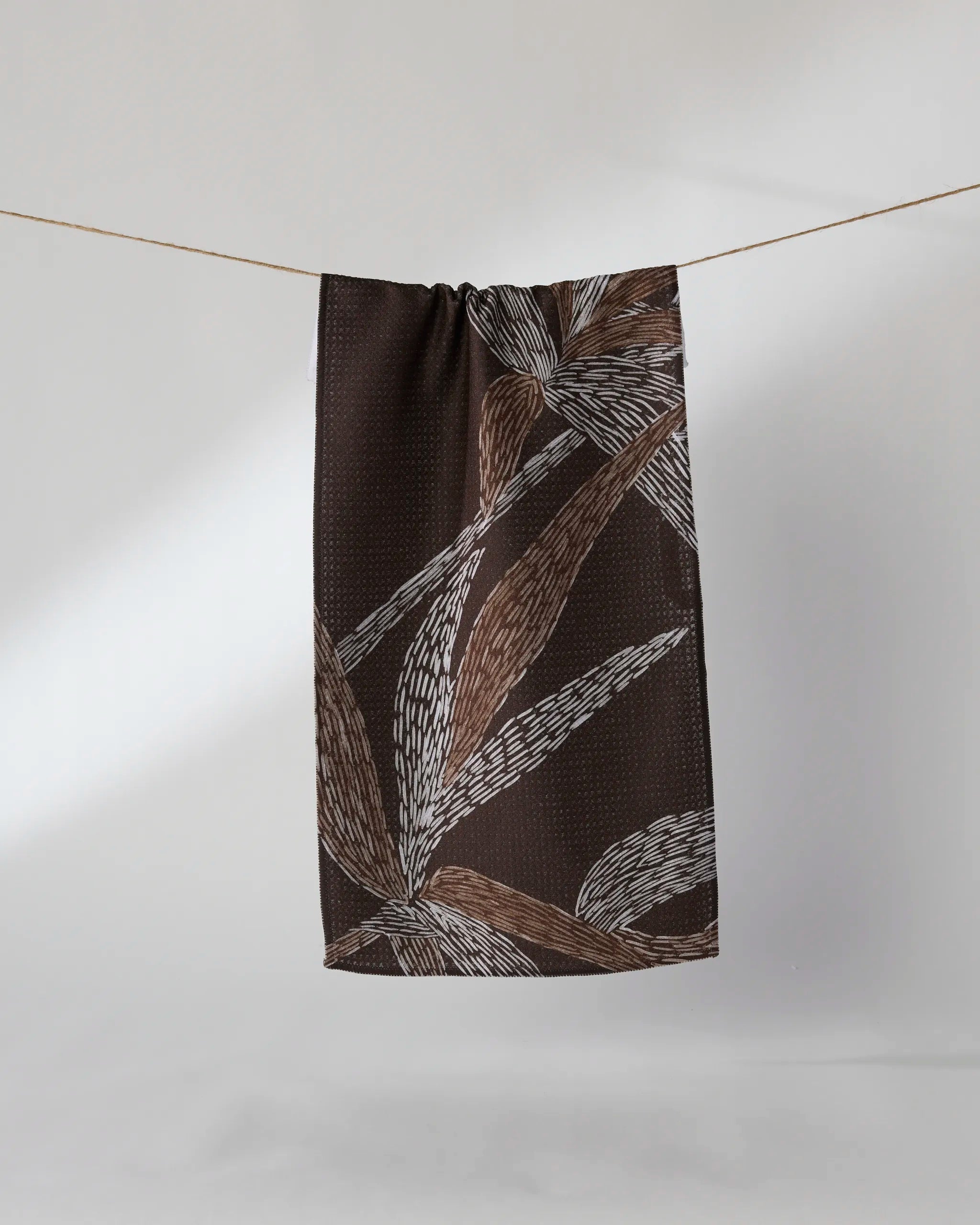 The Geometry "Midnight Wildflower" towel, featuring a dark brown hue with a white and beige abstract leaf pattern, hangs on a clothesline against a plain light background.