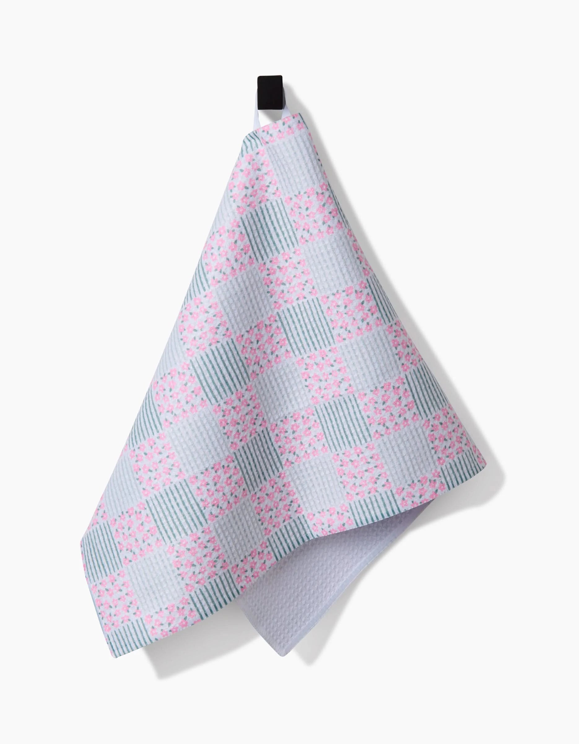 The Hufton Studio Mini Flowers and Stripes Check Pink and Green tea towel hangs from a black loop against a white background, featuring pastel pink and green checks crafted from Geoweave fabric for enhanced absorbency and durability.