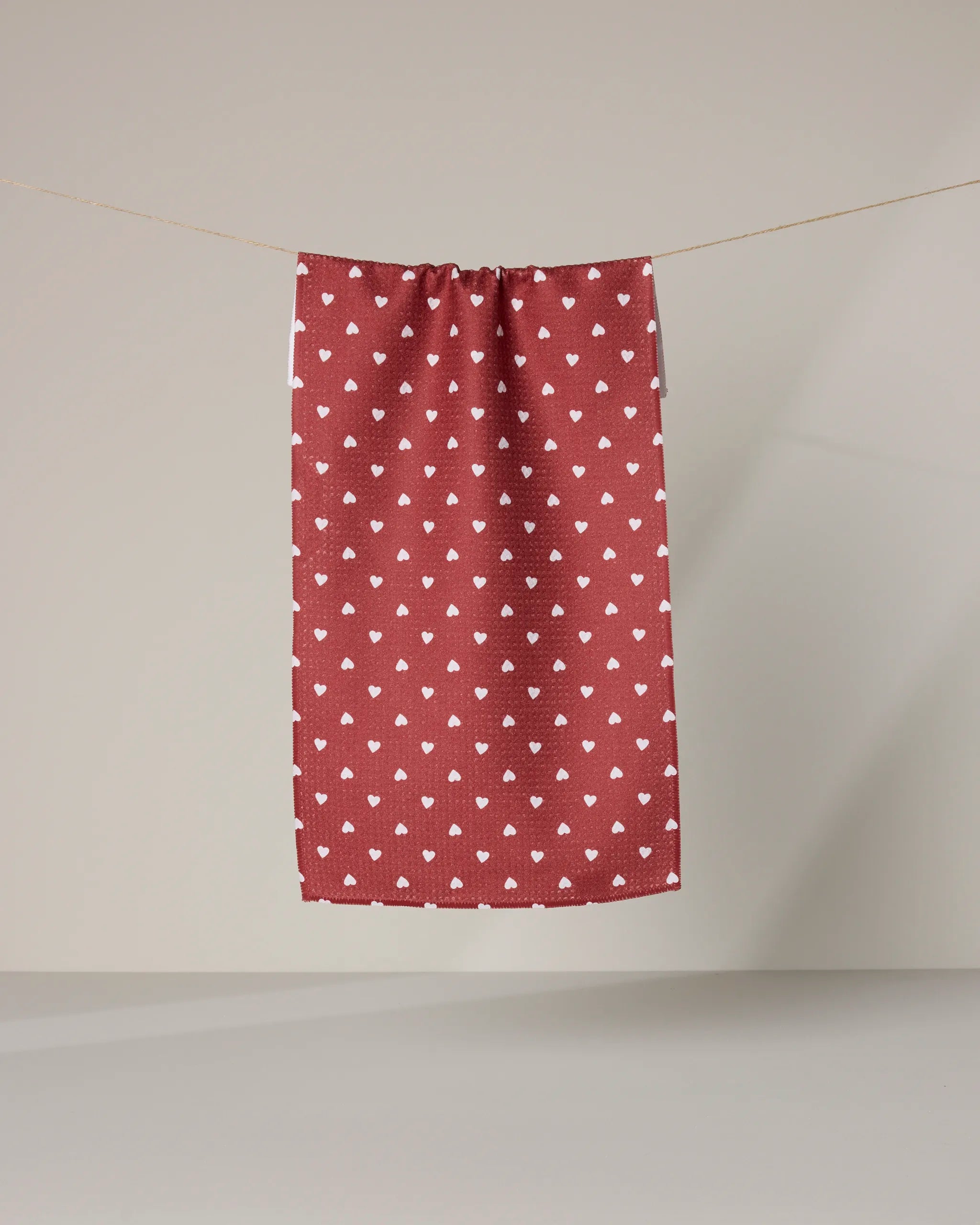 The Geometry Mini Heart bar towel, featuring a white heart pattern on red and made from recycled materials, hangs by a thin string against a plain light background.