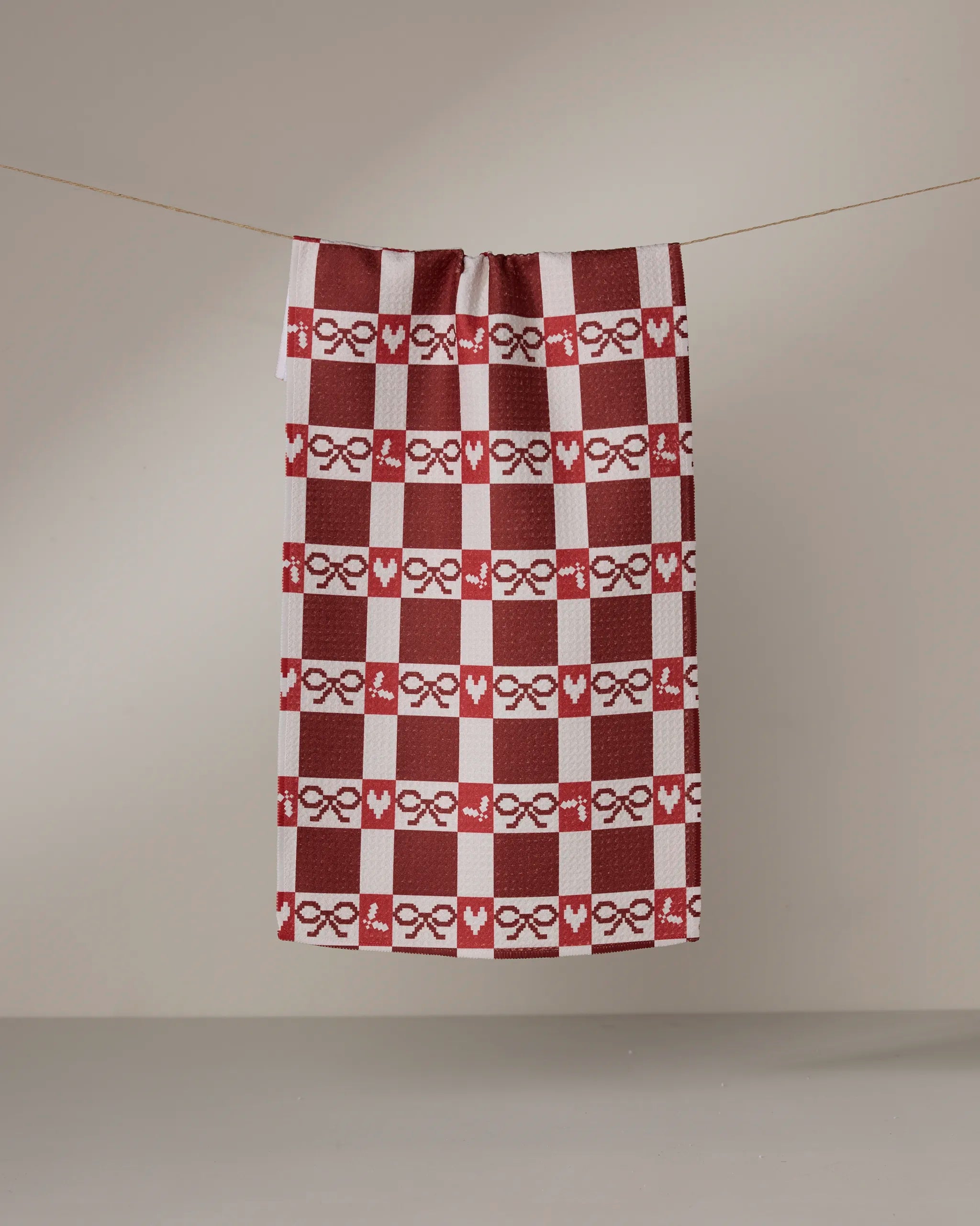 The Geometry Mistletoe Gingham bar towel, featuring red and white checks with heart and bow patterns and made from recycled materials, hangs from a thin line against a plain, neutral background.