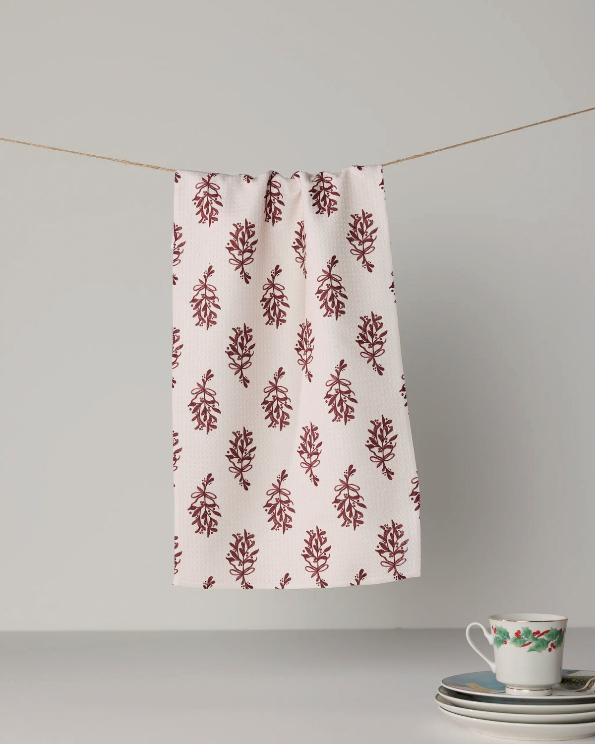 A Geometry “Mistletoe Sprigs” white cloth made from Geoweave™ fabric, featuring a maroon leaf pattern, hangs from a string. On the table below are stacked plates and a floral teacup, all set against a plain background.