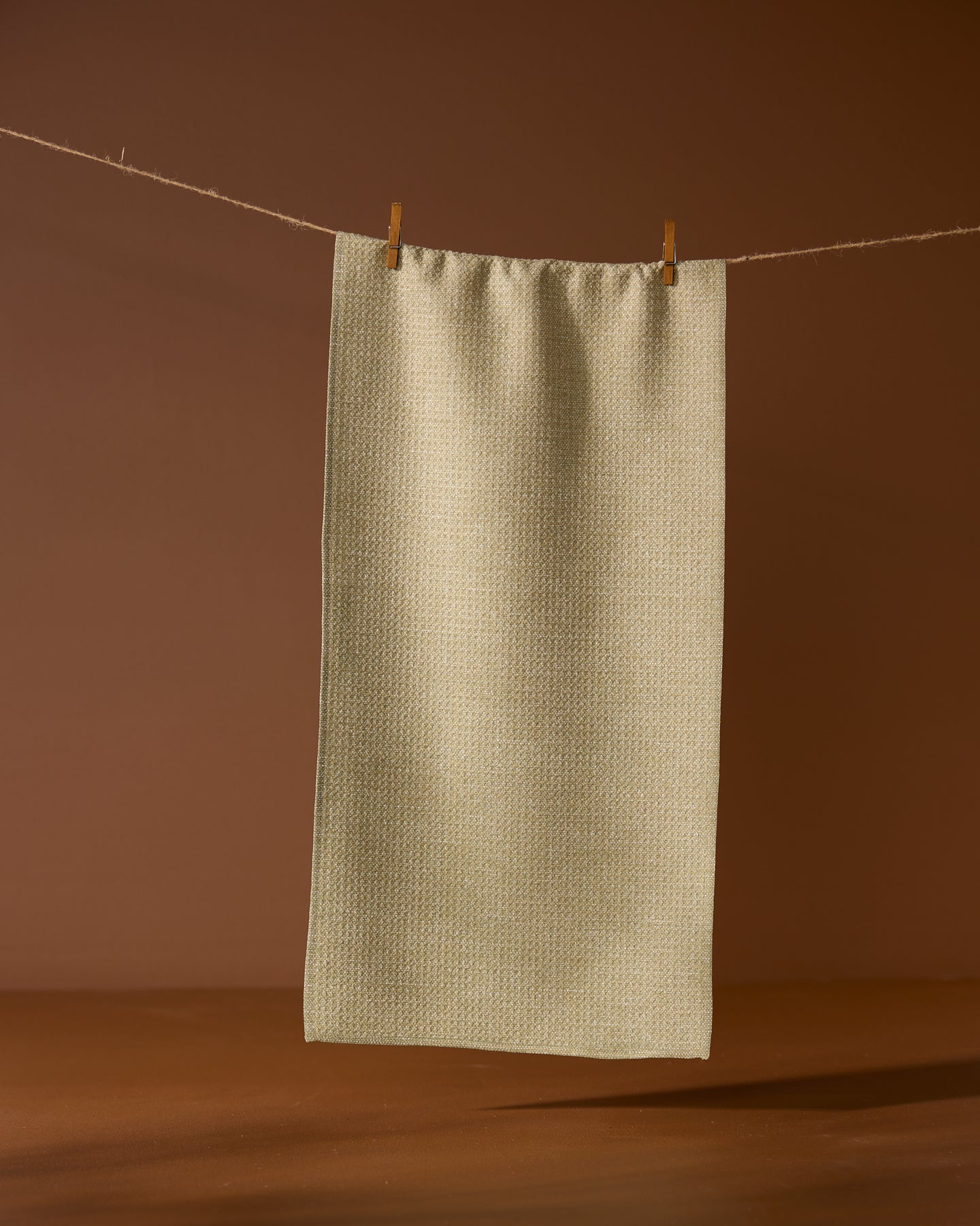 The Geometry Mosswood recycled kitchen towel in light beige hangs from a string with two wooden clothespins, set against a brown background.