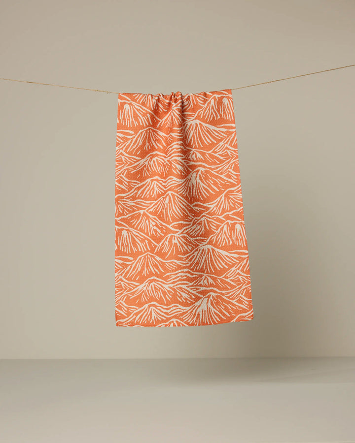The Geometry Mountain Range bar towel, featuring an orange and white Geoweave fabric with a mountain pattern, hangs on a string against a plain beige background.