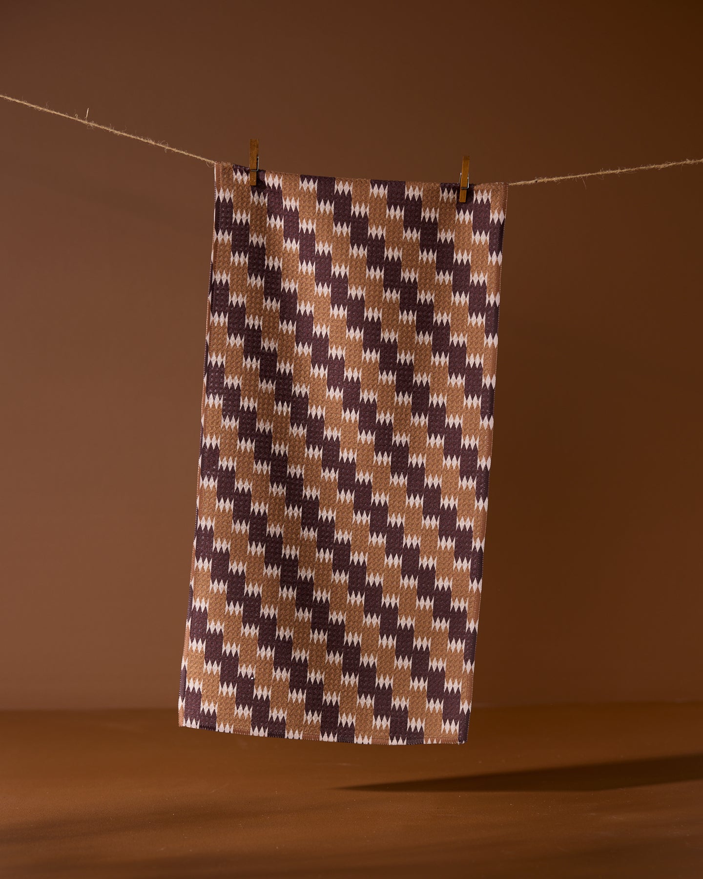 The Geometry Mountain Song bar towel features a brown, beige, and dark purple houndstooth pattern and hangs against a brown background. Made from recycled materials, it’s an eco-friendly and stylish choice.
